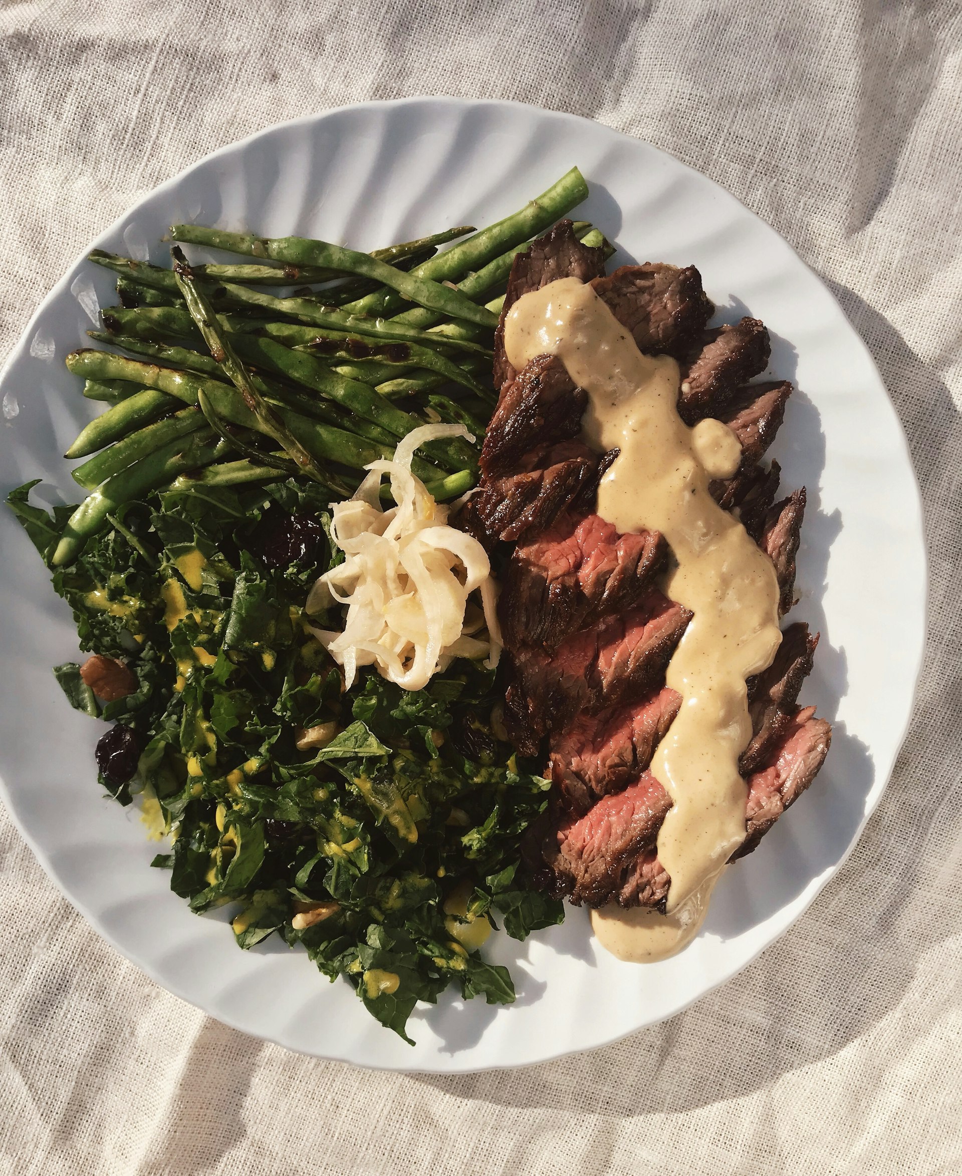 A white plate covered in thinly sliced strips of steak and a generous heap of greens is covered in a drizzle of caramelized brunost sauce against a linen backdrop