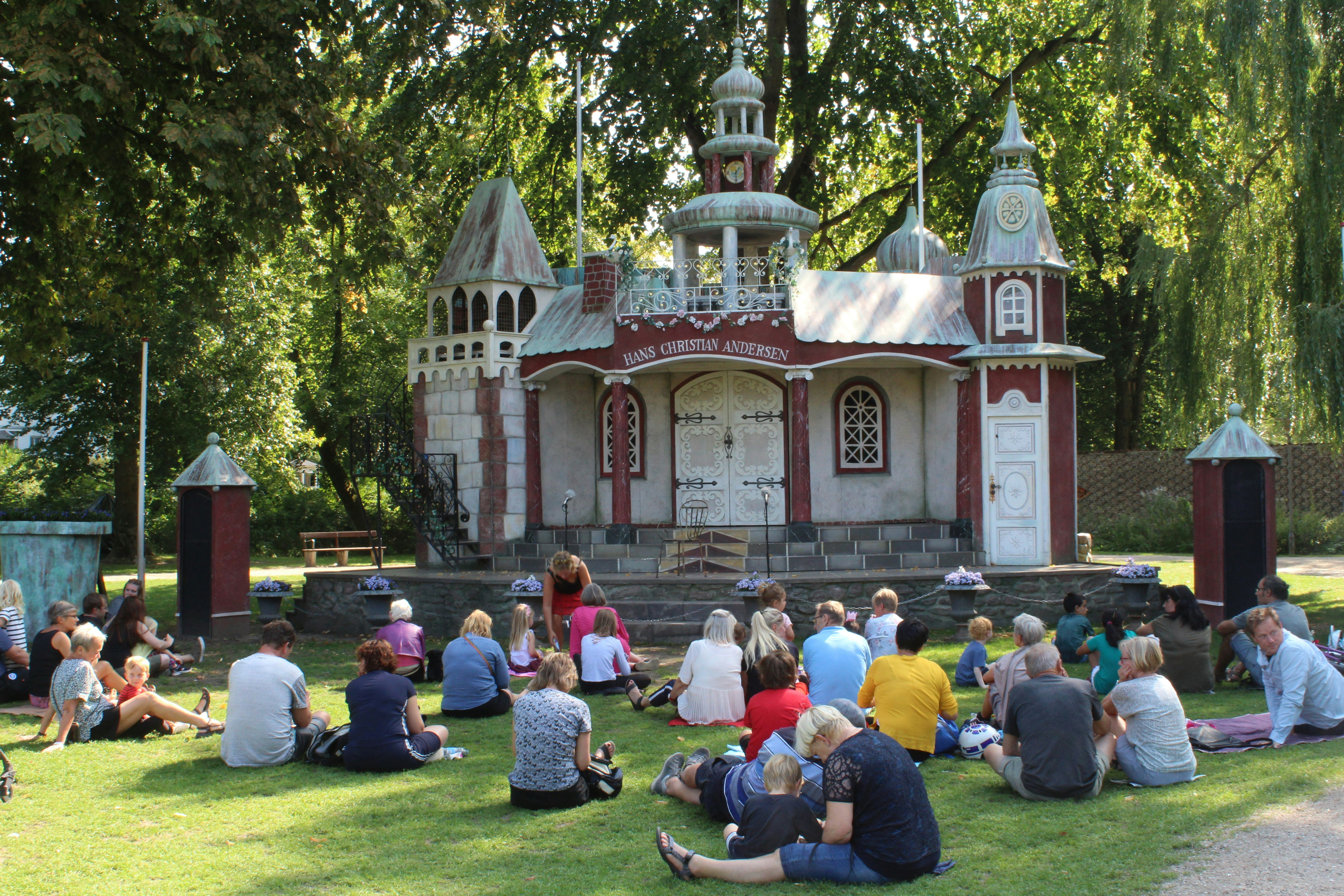 Families are sitting on the grass in a park on Eventyrhaven island, facing the stage of the miniature Hans Christian Andersen castle.