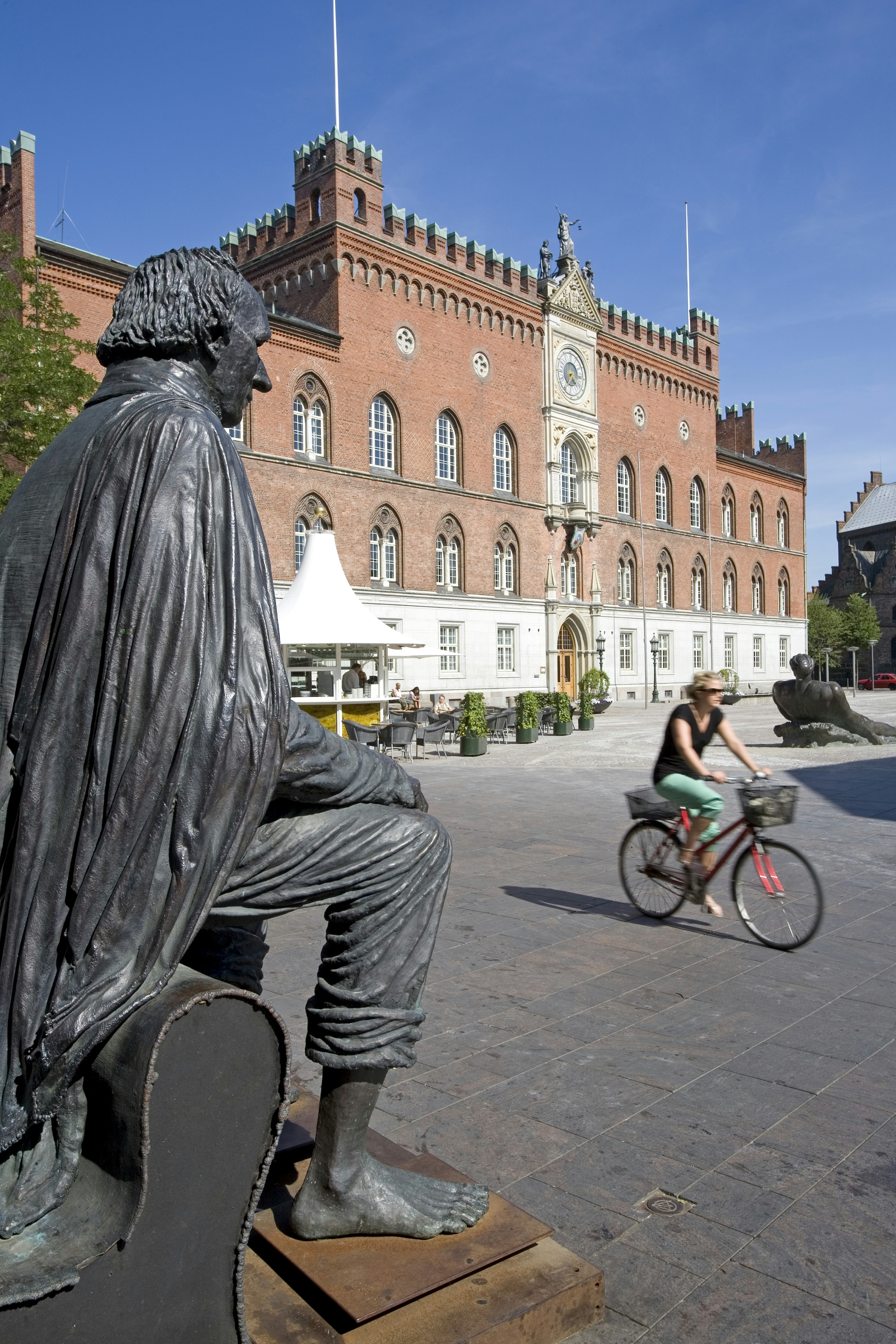 A seated statue of Hans Christian Andersen on a large public square in Odense, Denmark; a woman is cycling across the frame in front of the statue.