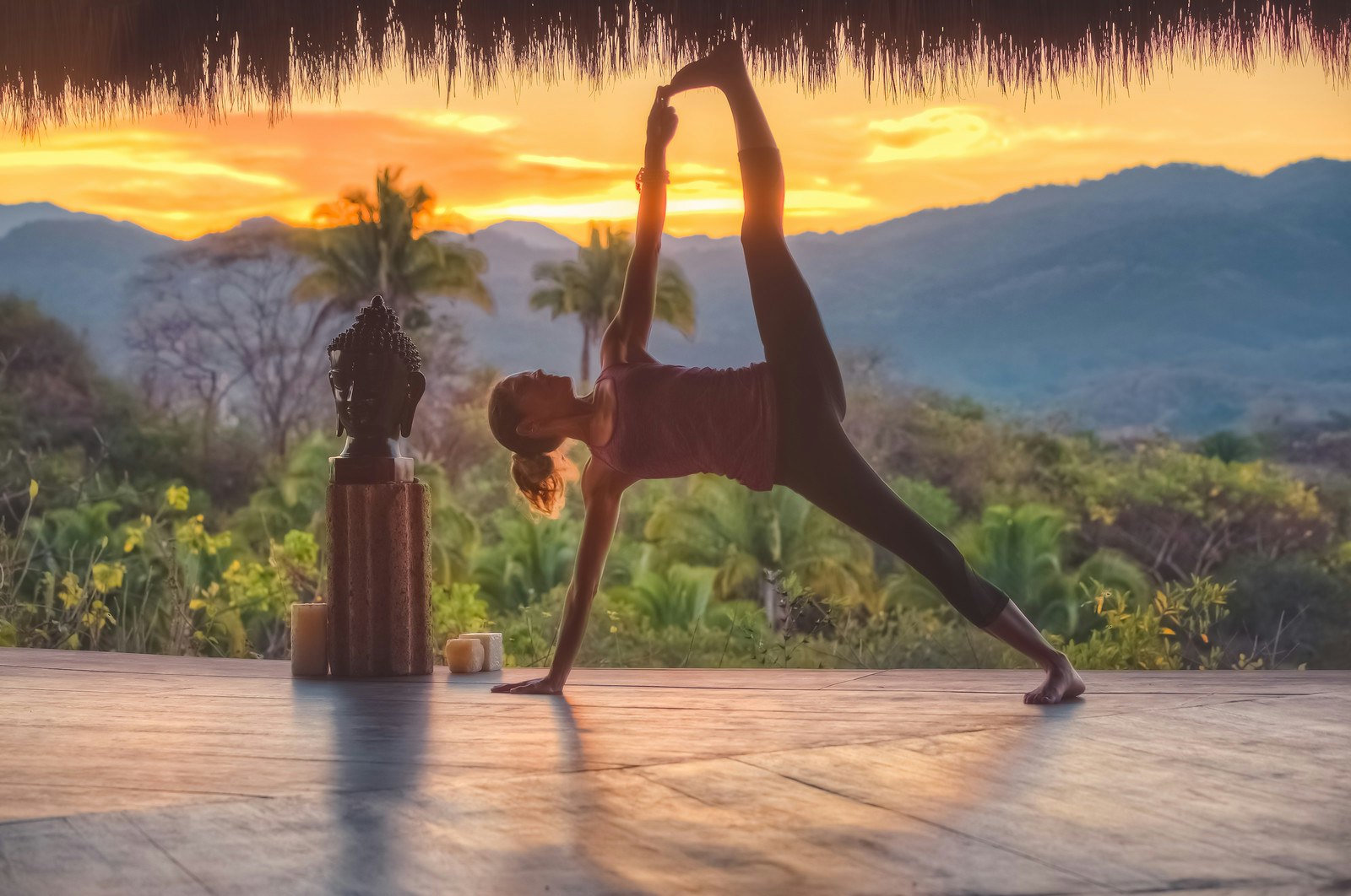 A woman is in a challenging yoga pose at luxury retreat. She is in an open-air yoga studio; behind her are lush tropical plants and trees, and in the distance are mountains with the sun setting behind.