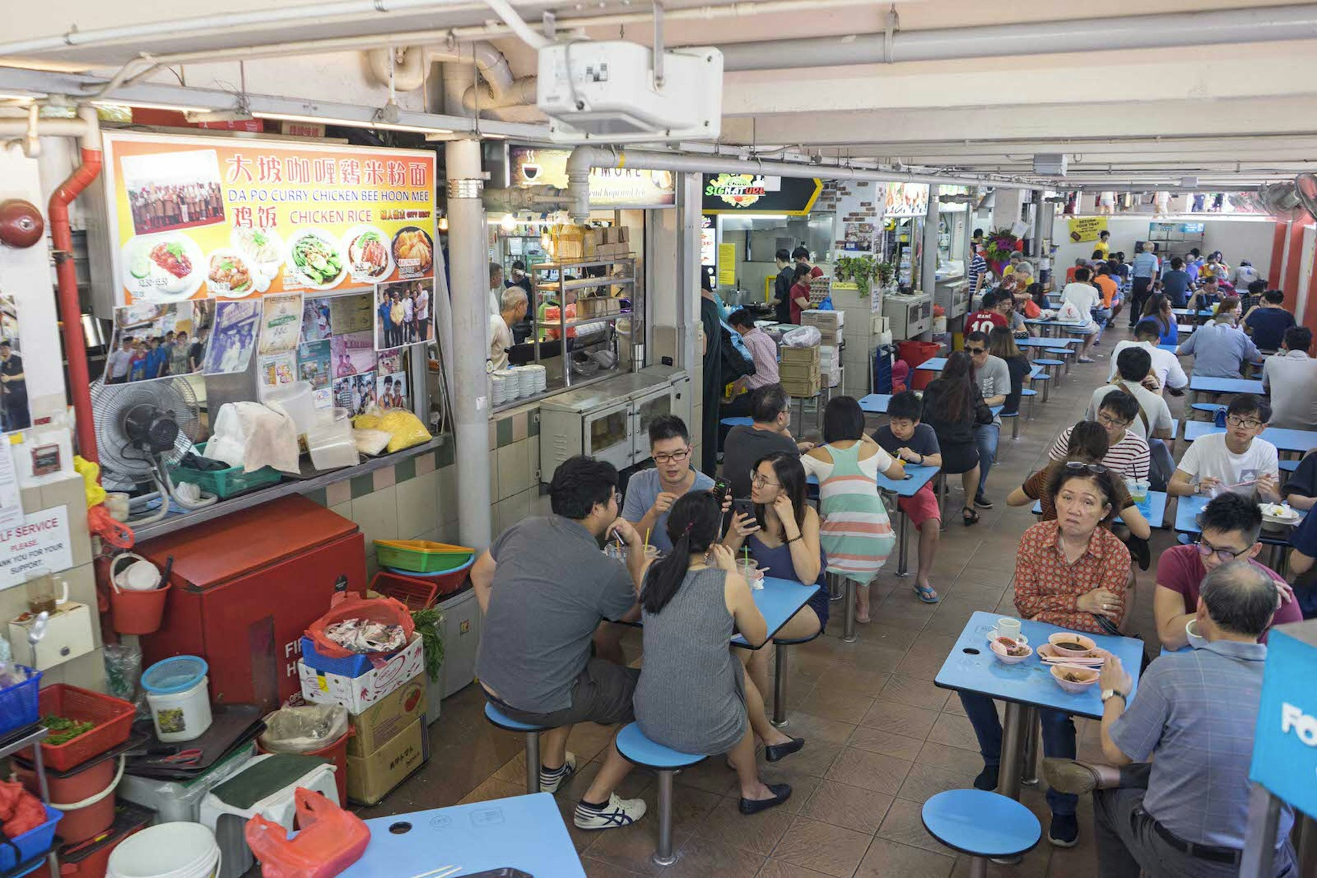 A busy hawker centre in Singapore. There are small blue tables with matching blue stools, almost every table is occupied by groups. Along the left side of the dining area there are various food stalls.
