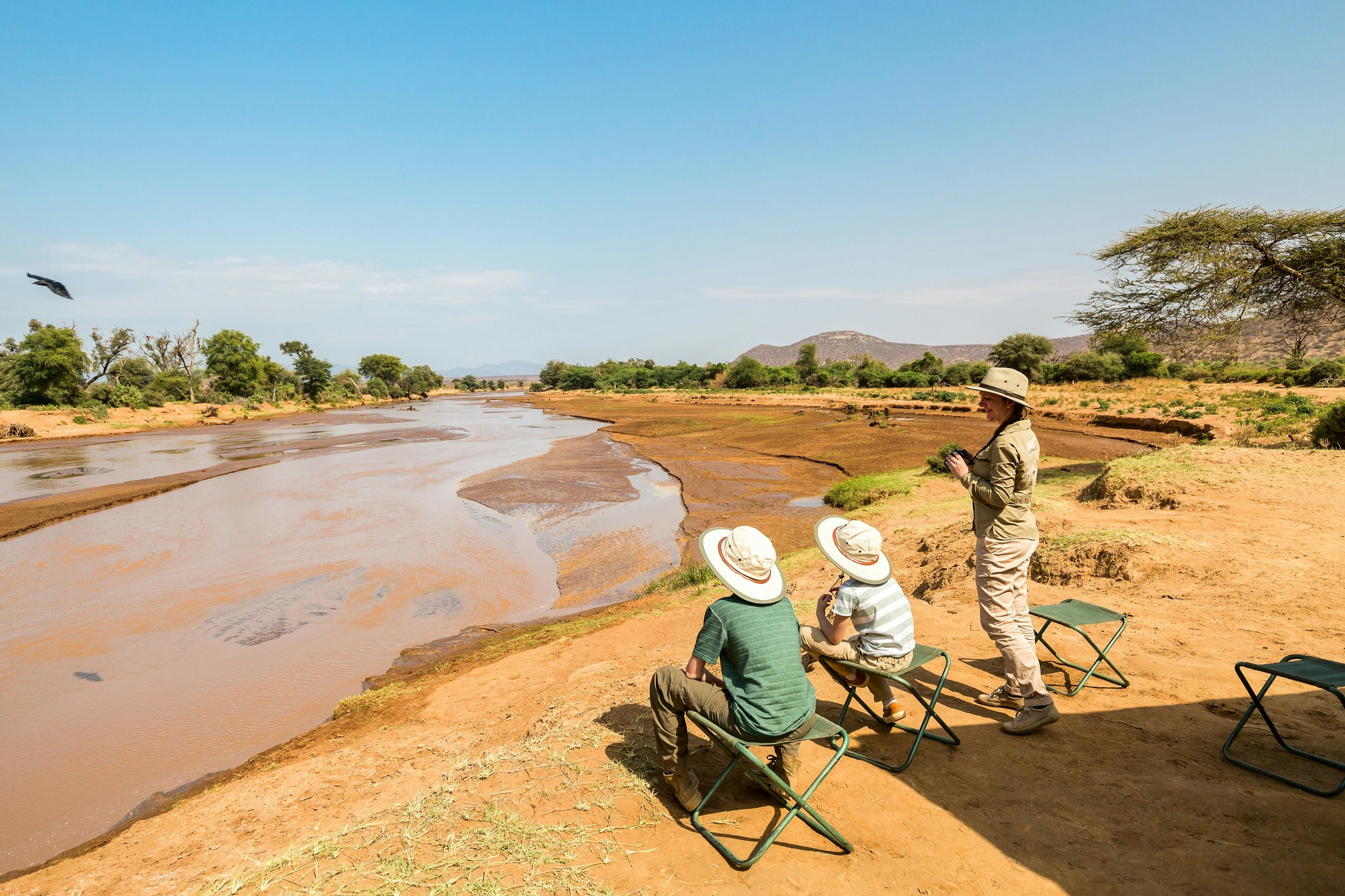 A mother stands next to her two kids (both on folding camping chairs) next to the muddy Ewaso Nyiro River; behind them is the shadow of their safari tent.