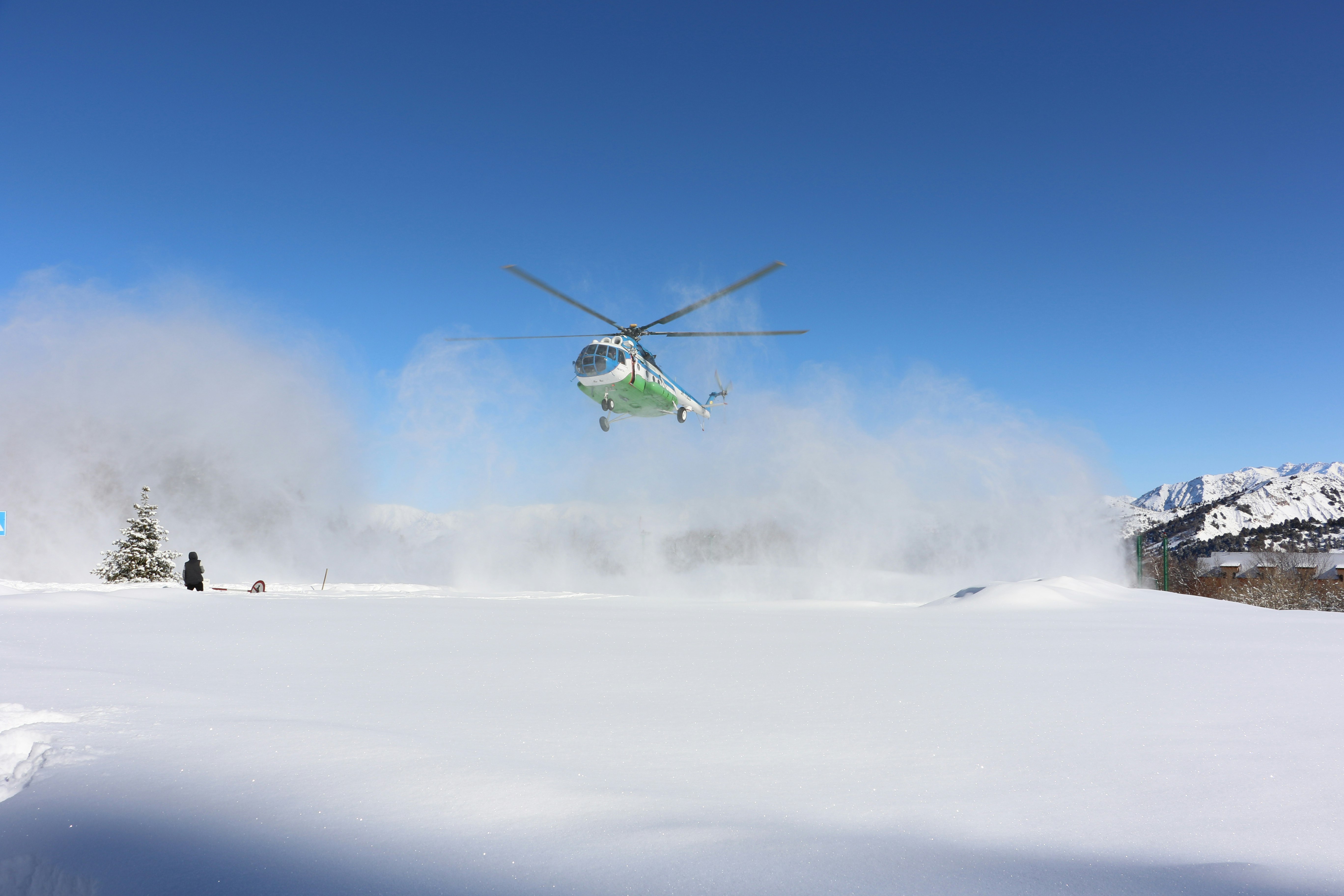 A Soviet Mi-8MTB helicopter takes off from a helipad. The ground is covered in snow and the sky is bright blue behind the white helicopter.