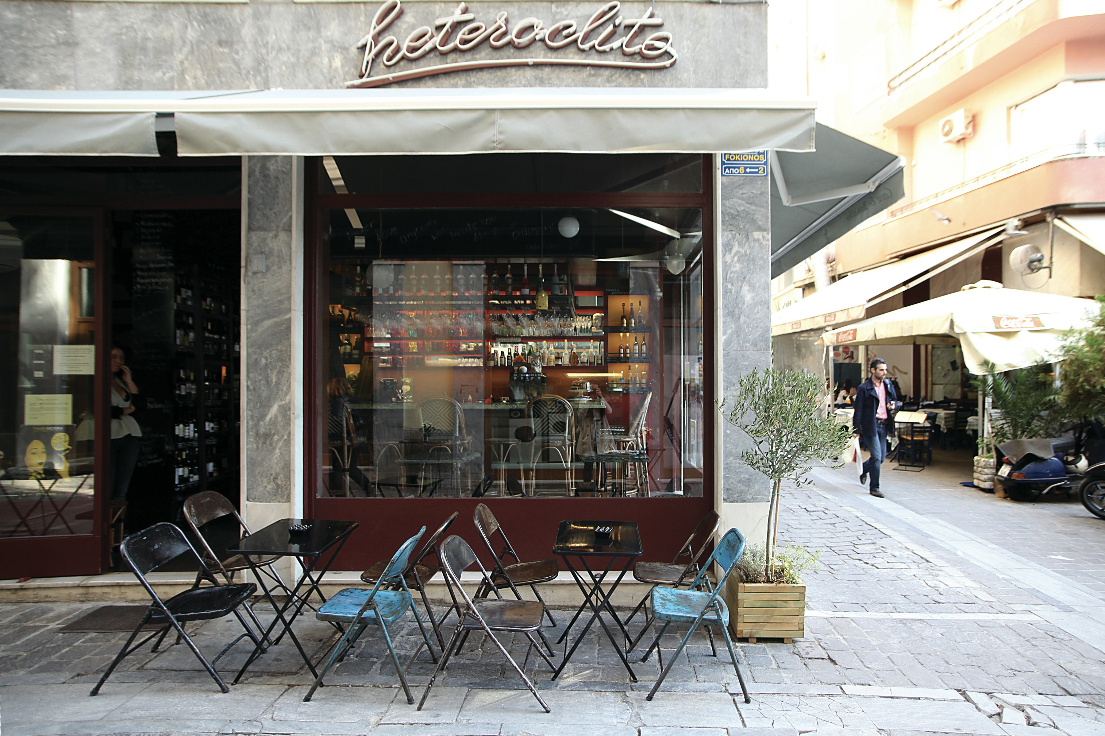 The silver exterior of the Hertoclito restaurnt in the Syntagma and Plaka neighborhood has blue and black cafe chairs out front and a small tree growing in a wooden planter box. Through the window you can see beautifully lit bottles behind the bar in dark, inviting tones. Outside, an Athenian man in jeans and a black jacket walks down the cobbled street