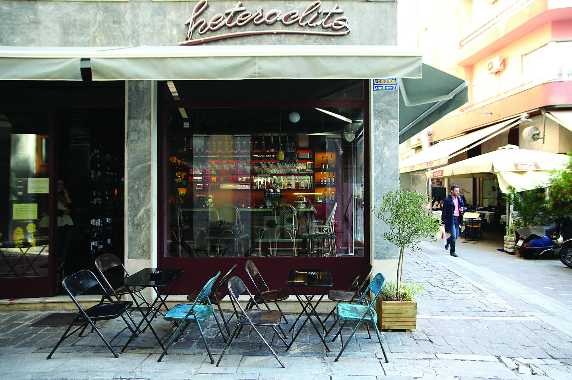 The silver exterior of the Hertoclito restaurnt in the Syntagma and Plaka neighborhood has blue and black cafe chairs out front and a small tree growing in a wooden planter box. Through the window you can see beautifully lit bottles behind the bar in dark, inviting tones. Outside, an Athenian man in jeans and a black jacket walks down the cobbled street