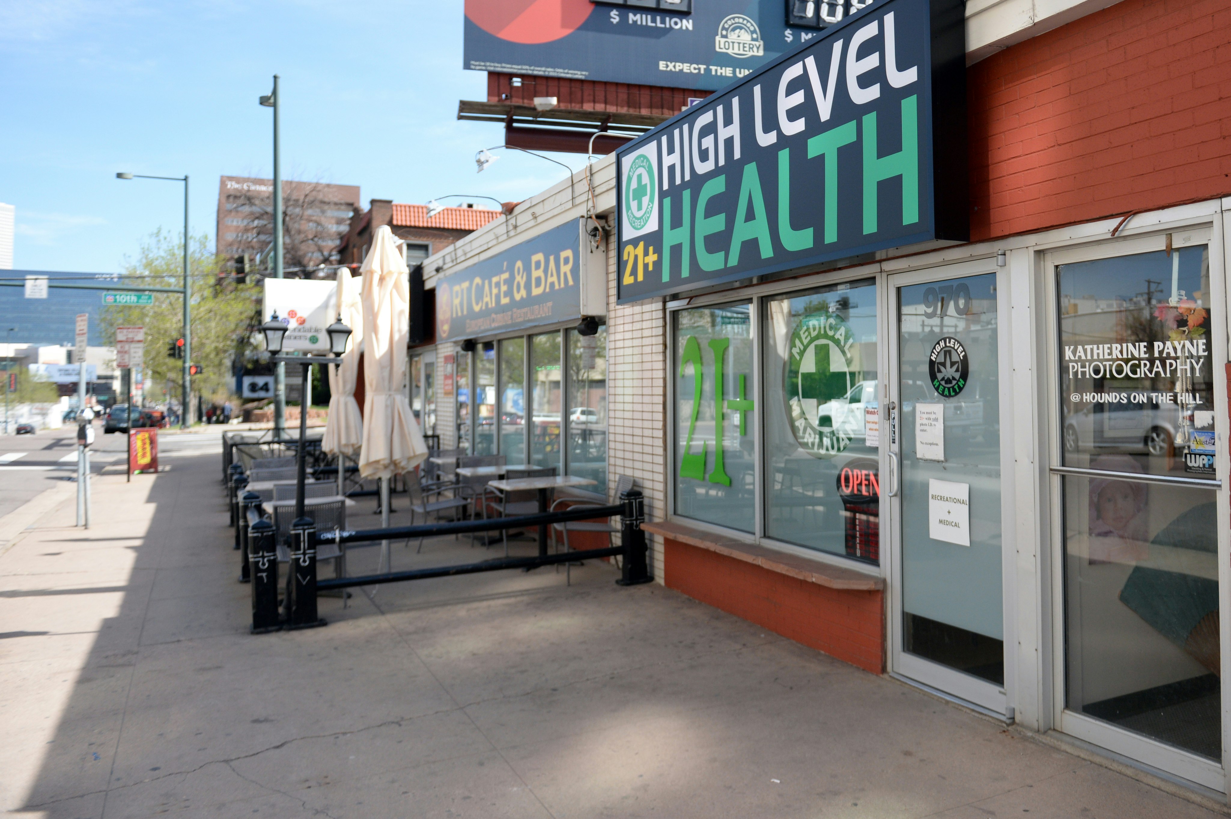 The exterior of High Level Health dispensary in Denver, Colorado has red brick around glass windows that are obscured by sheets of white paper. A large green sign reads 21+, while another shows the Medical Marijuana logo. Right next door is a cafe and bar with outdoor patio seating