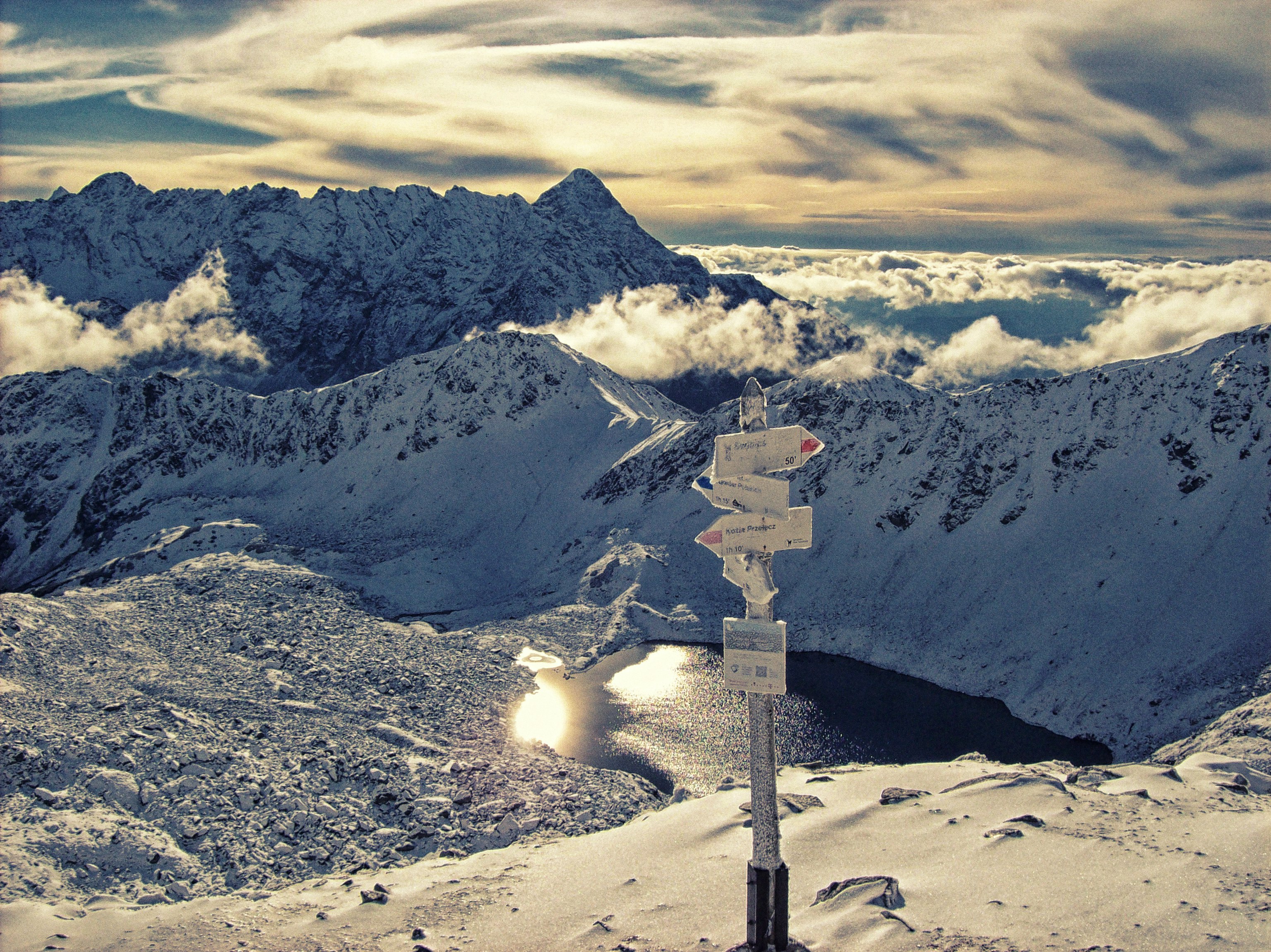 A mountain scene looking down towards a lake in Poland's High Tatras; the terrain is entirely covered in snow, and there's a wooden post with several destination markers on it. 