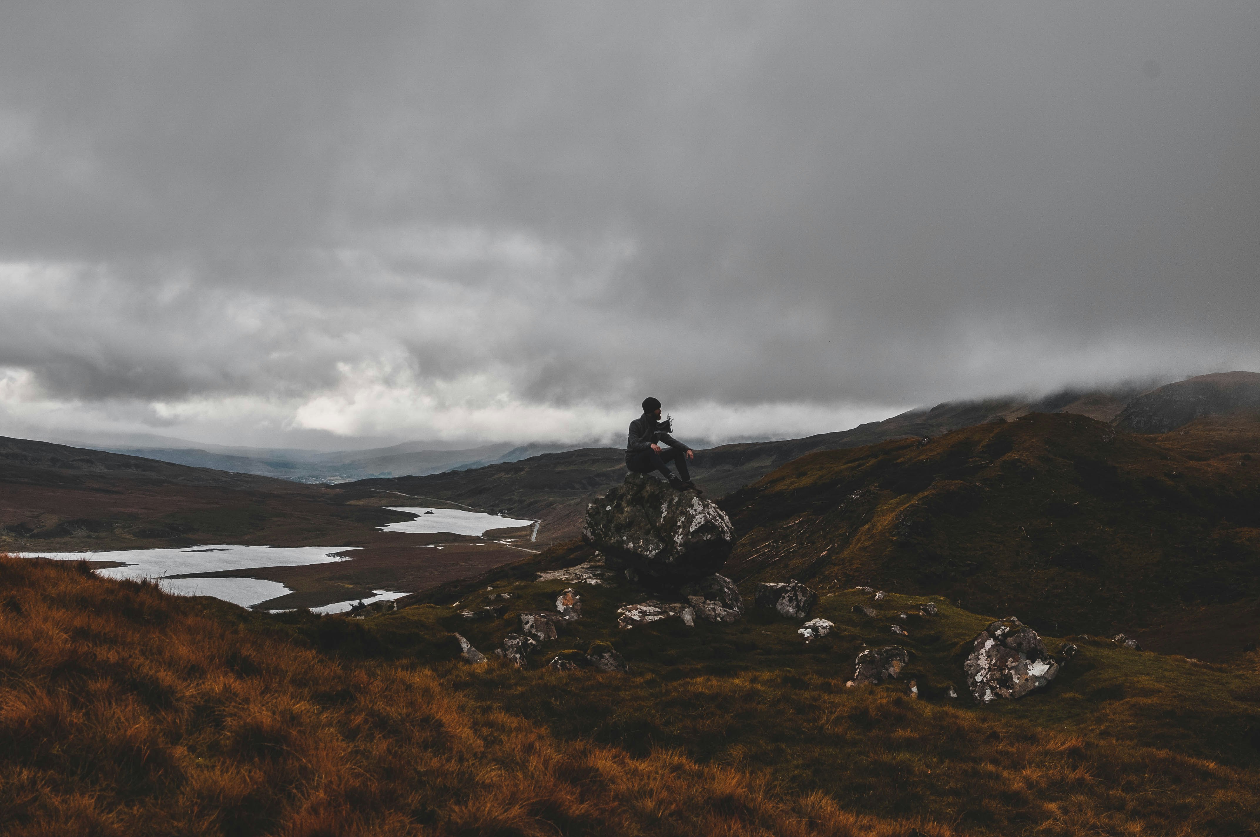 A man sits on a rock in a deserted Highland landscape. In the background is a loch and mountains covered by fog.