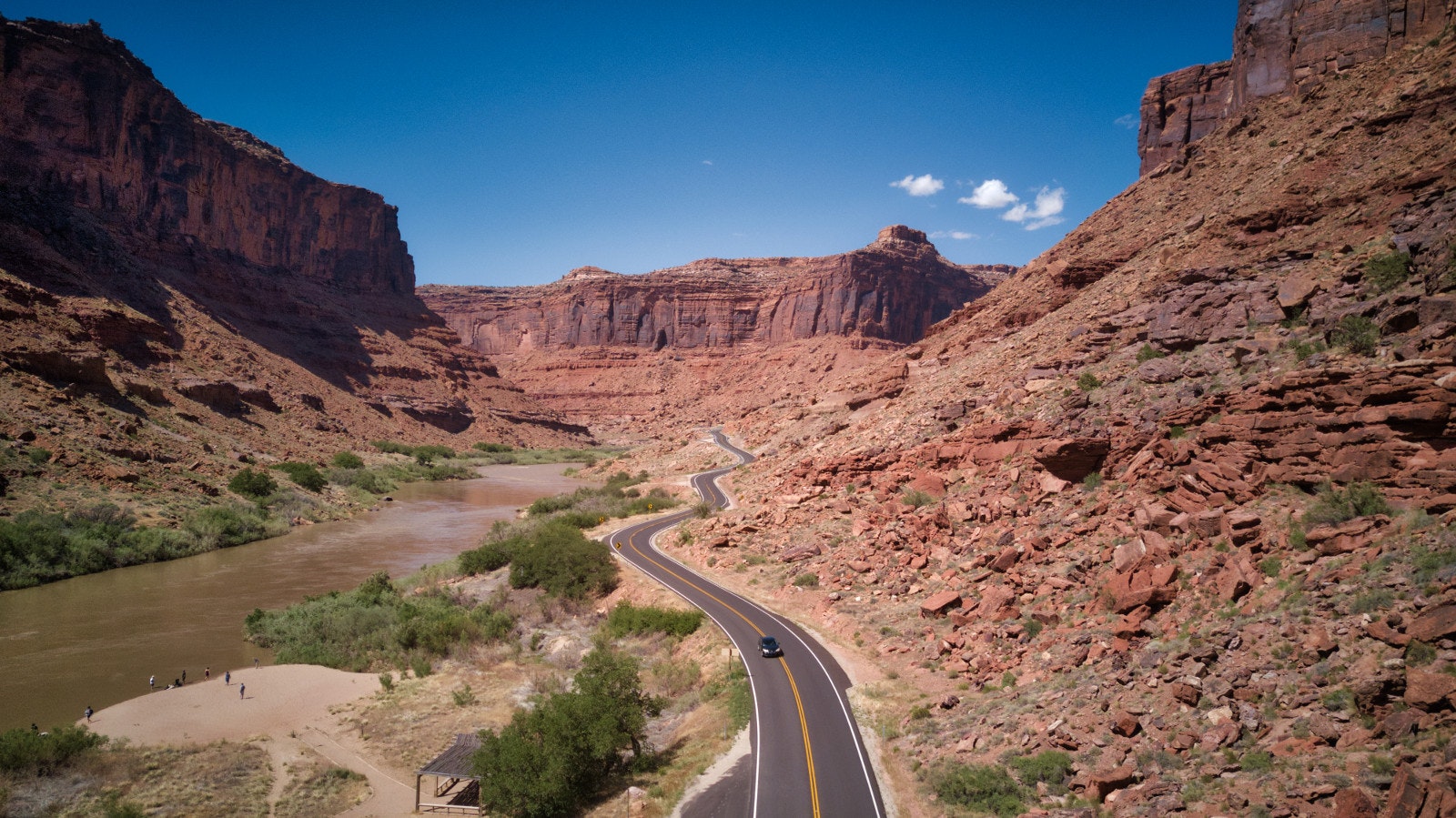 A road winds through a large red canyon, with one car on it. On the left is a river with some people on the edge.