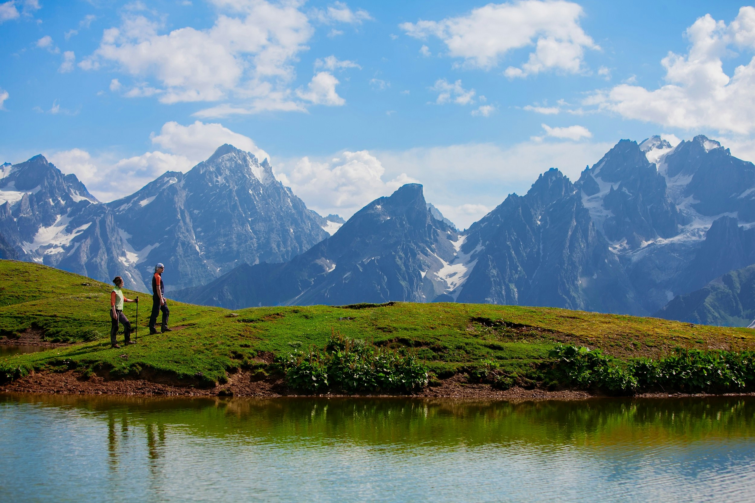 Two people walk beside a lake. In the distance is a huge range of pointed mountains.