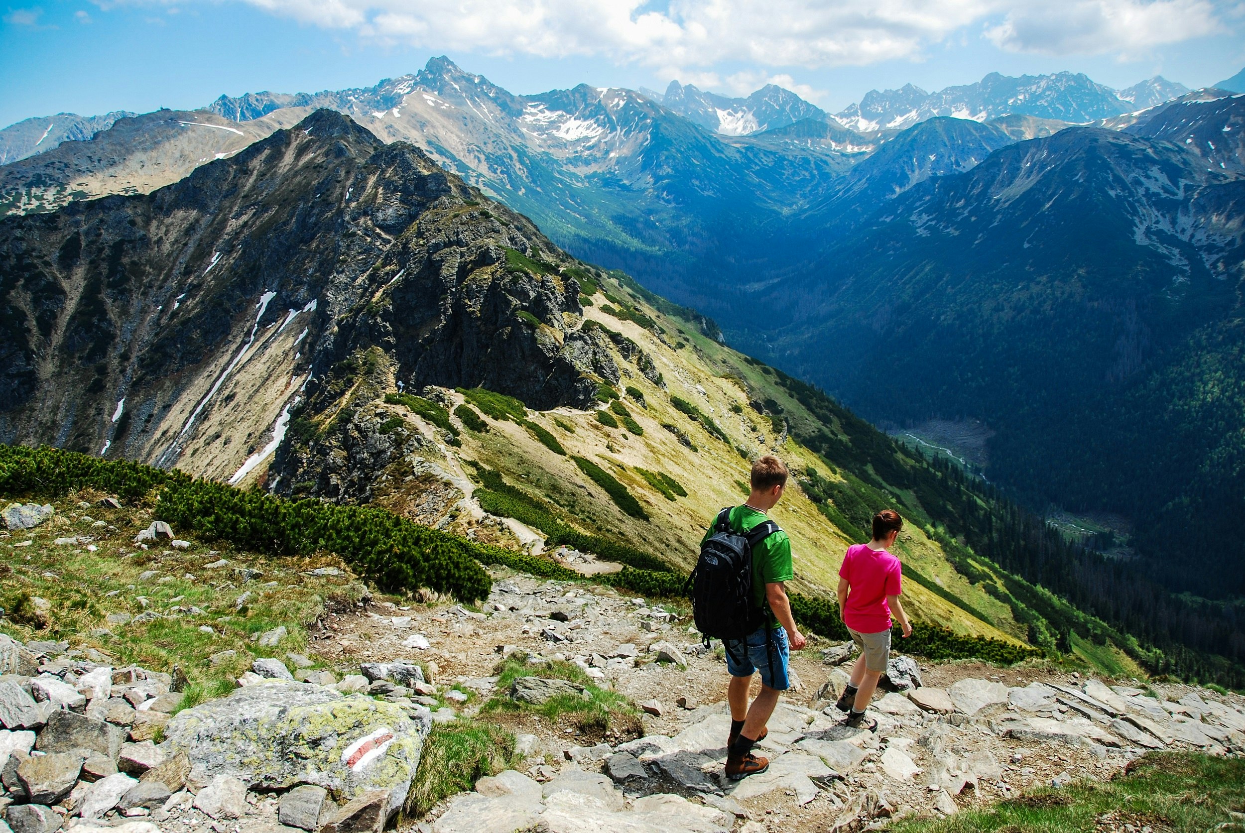 Two hikers dressed in summer clothes follow a rocky path down a hillside. A mountain range stretches out in the distance