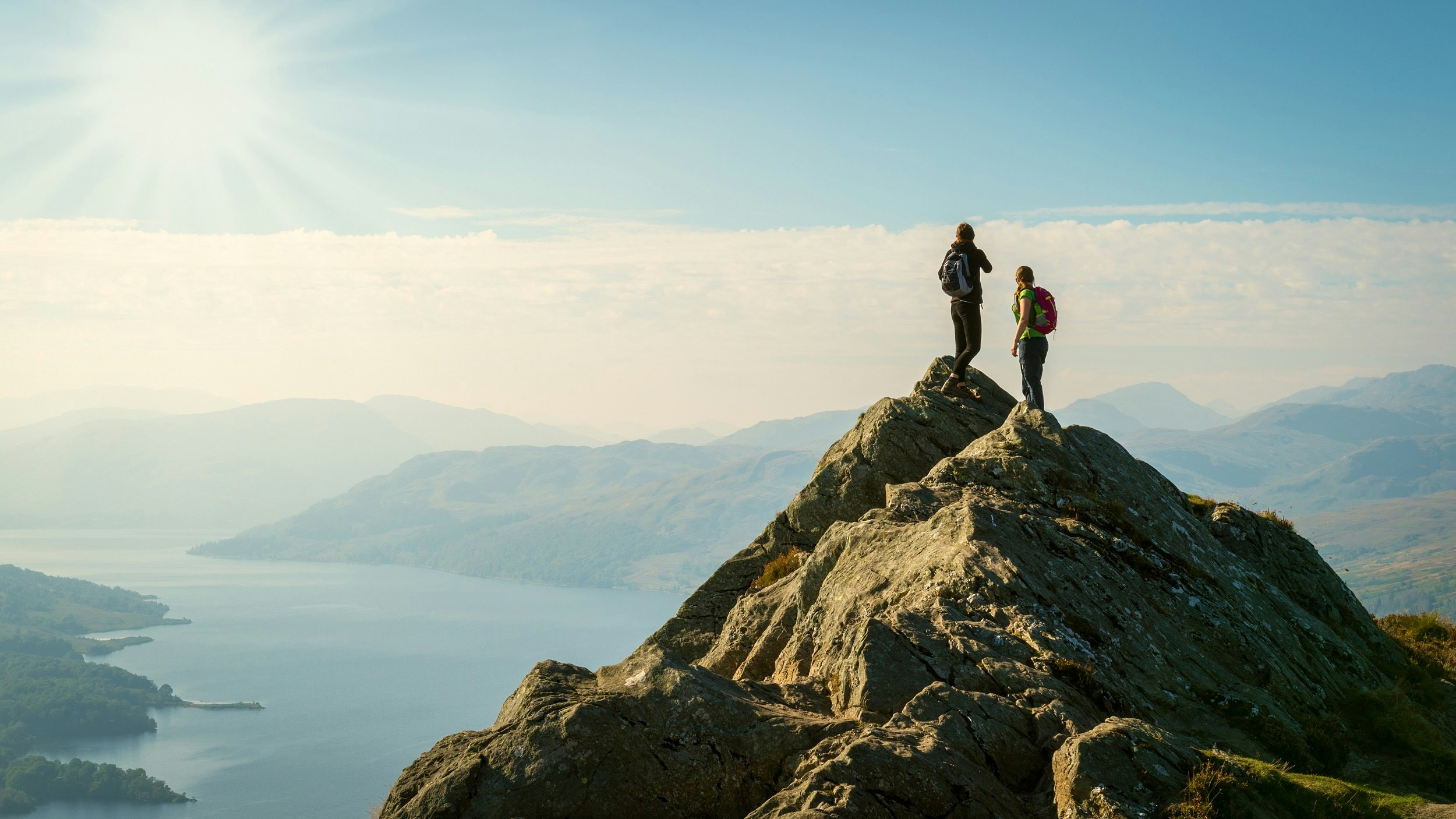 Two hikers wearing backpacks stand on a peak looking out over a loch in Scotland