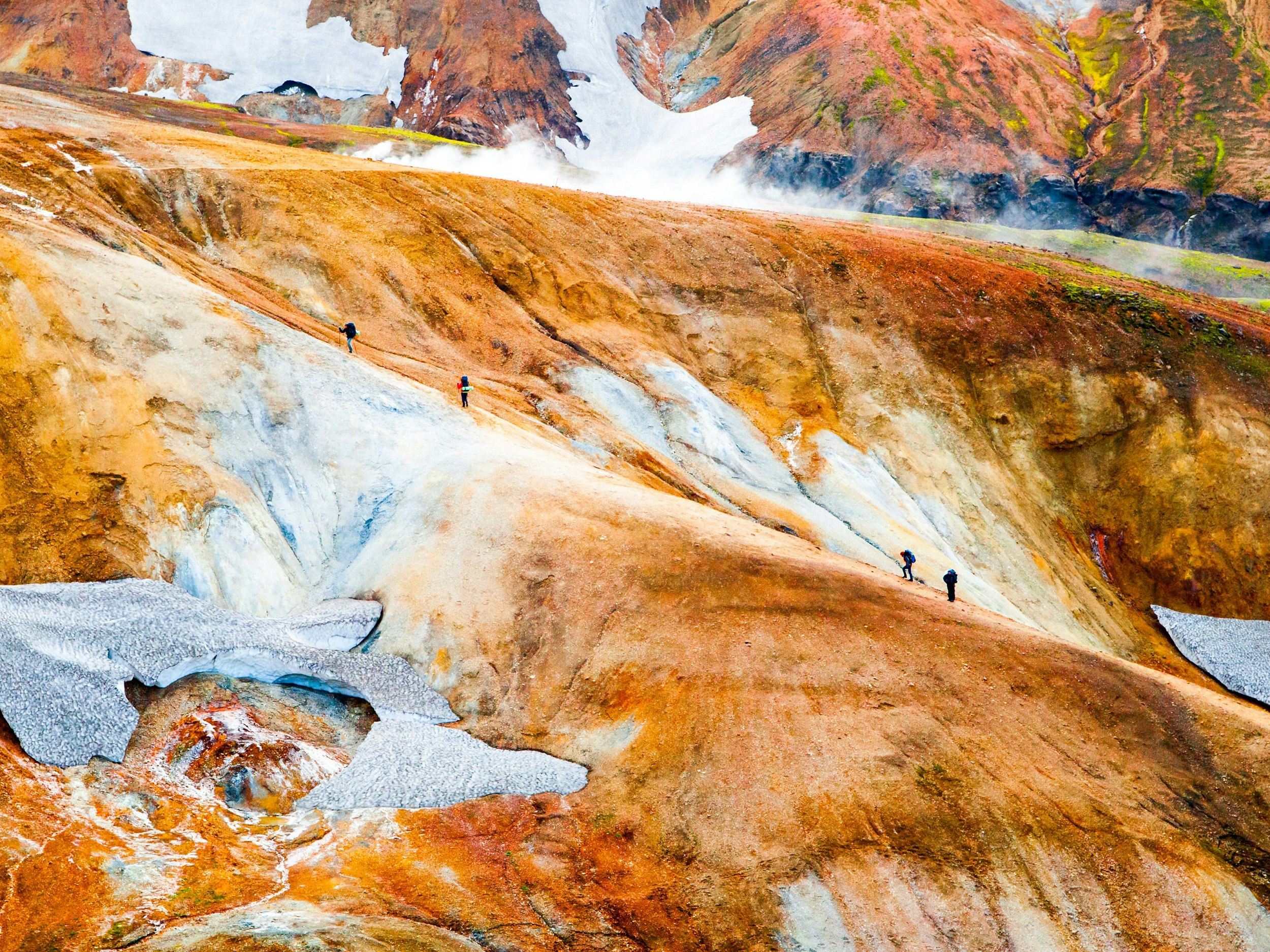 Four trekkers, in two groups of two, walk up a rusty coloured ridge that is flanked by small remnants of glaciers.