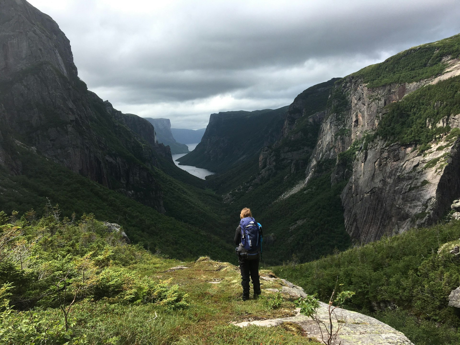 A woman wearing a backpack stands at the head of a spectacular fjord with her back to the camera in Gros Mourne National Park, gazing at the view ahead.