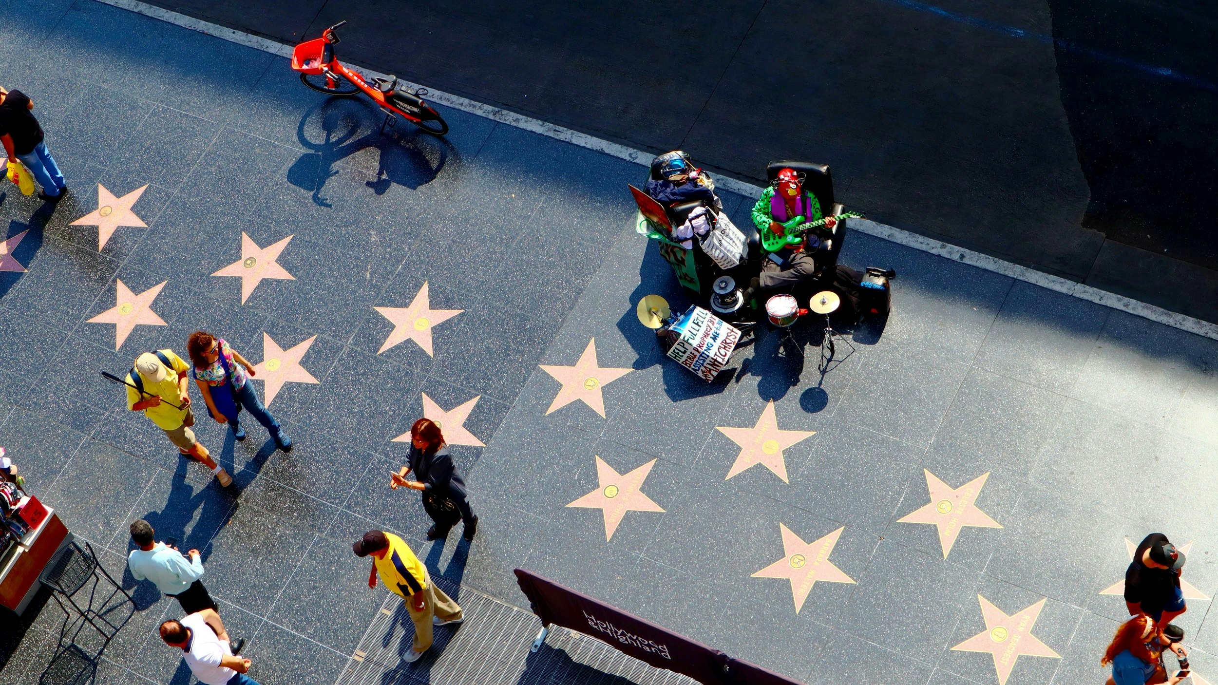 This image is looking down on a section of dark grey pavement with numerous large stars bedded into it, each of which carries the name of a famous person in show business; people walk to and fro.  