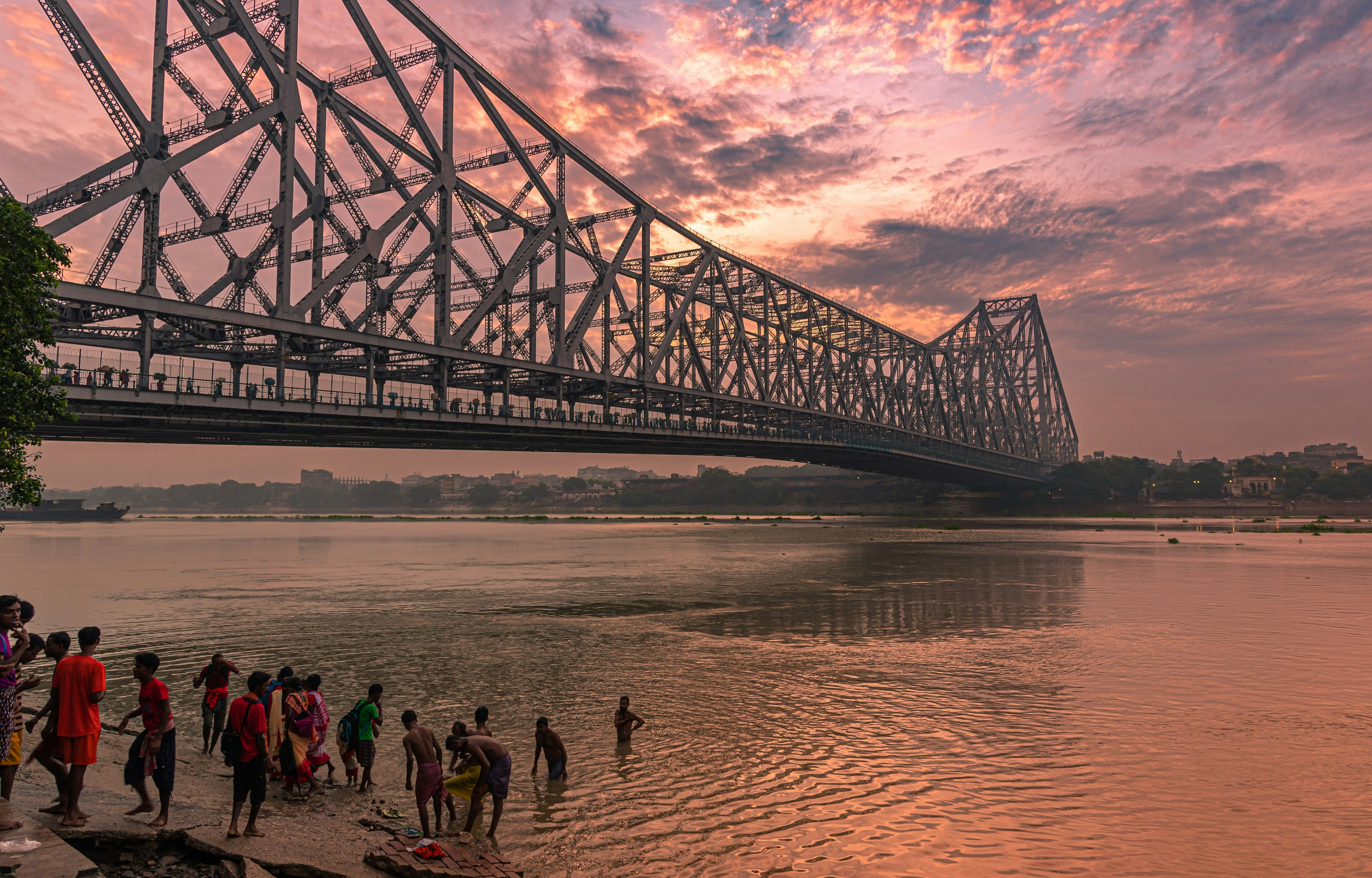 A group of children bathe in the Hooghly River next to one of Kolkata's large bridges in the early evening light.