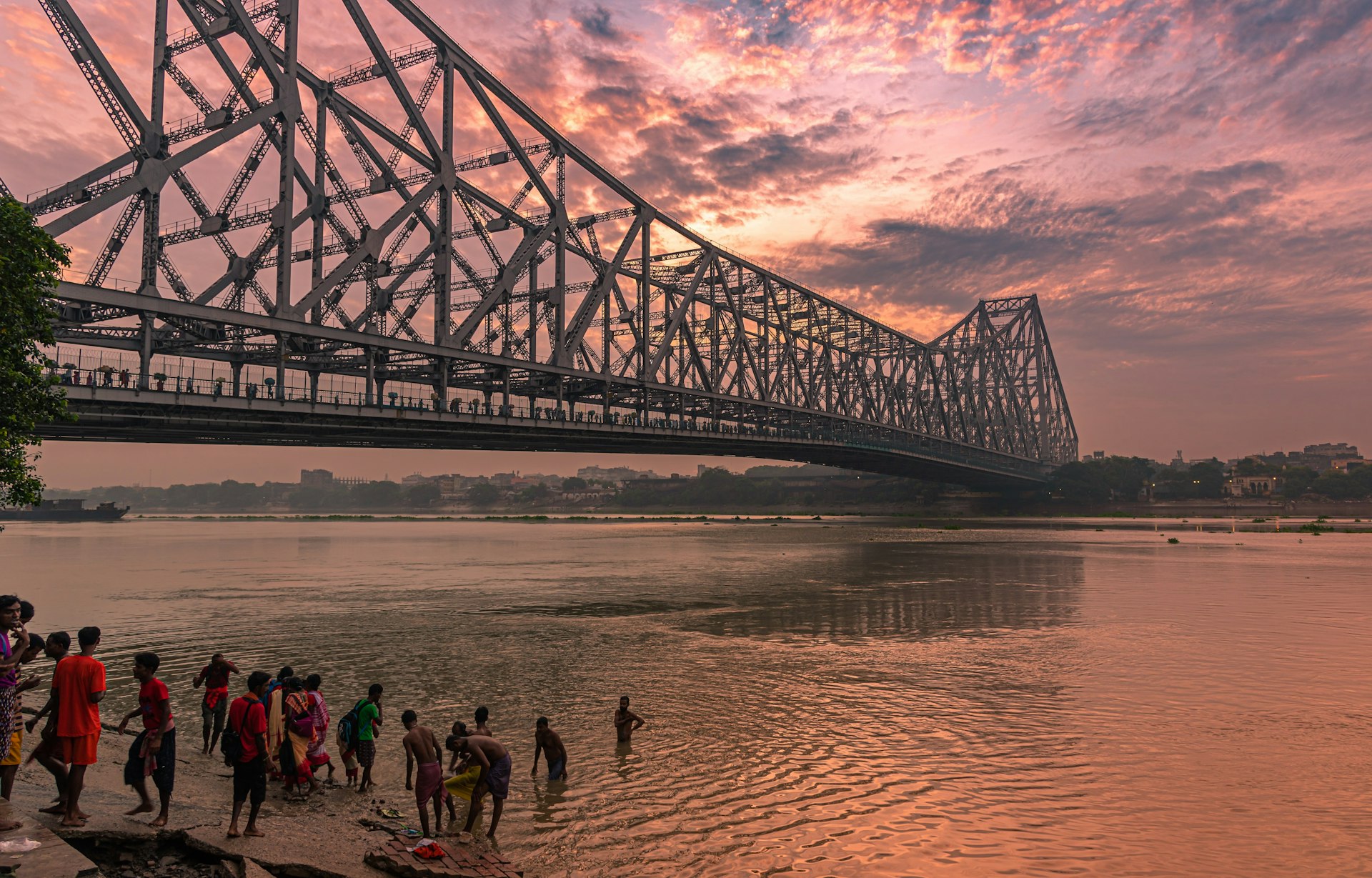 A group of children bathe in the Hooghly River next to one of Kolkata's large bridges in the early evening light.