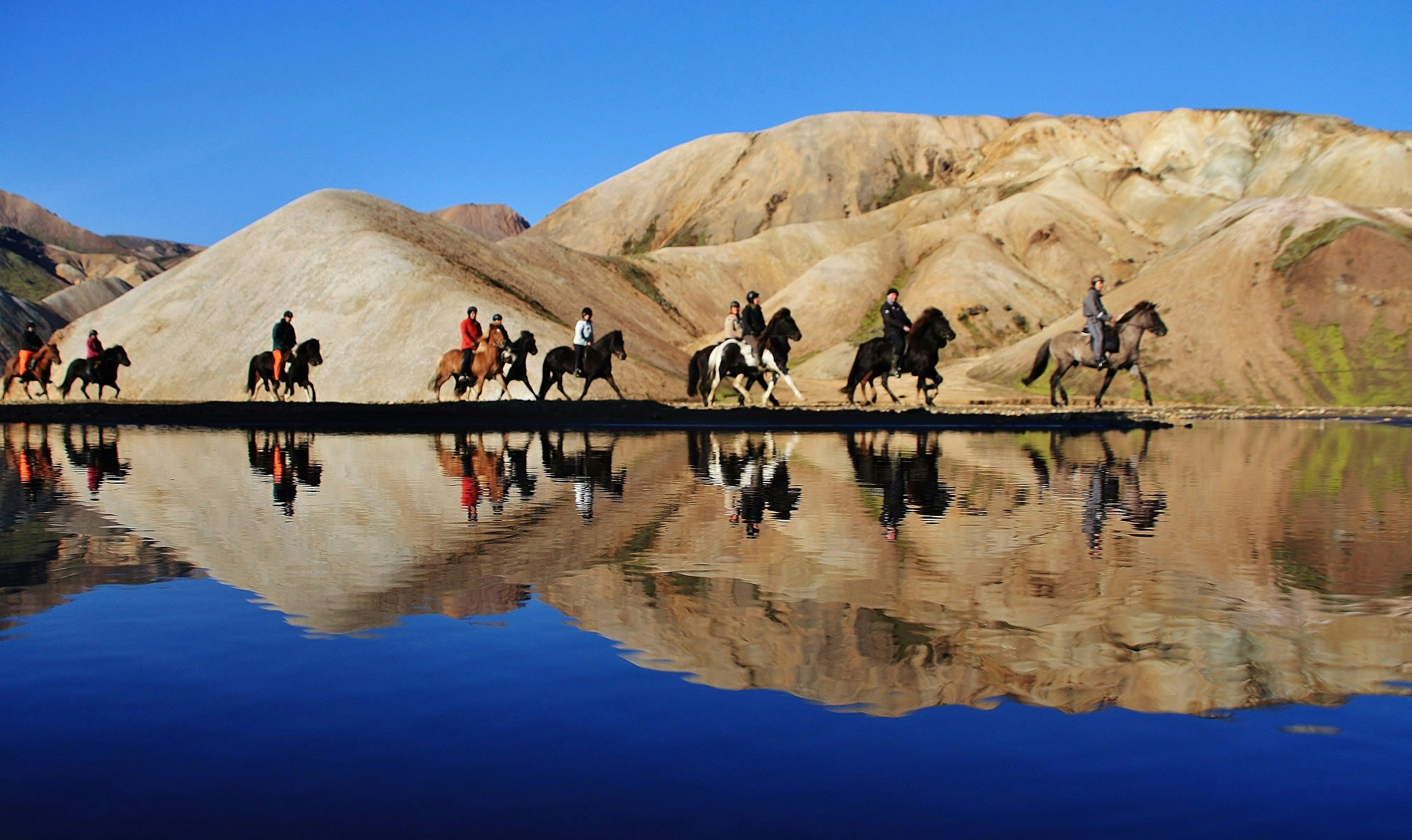 A dozen people on horseback make their way along a mirror-calm lake, with their reflections showing perfectly; behind them are some barren, tan-coloured hills.