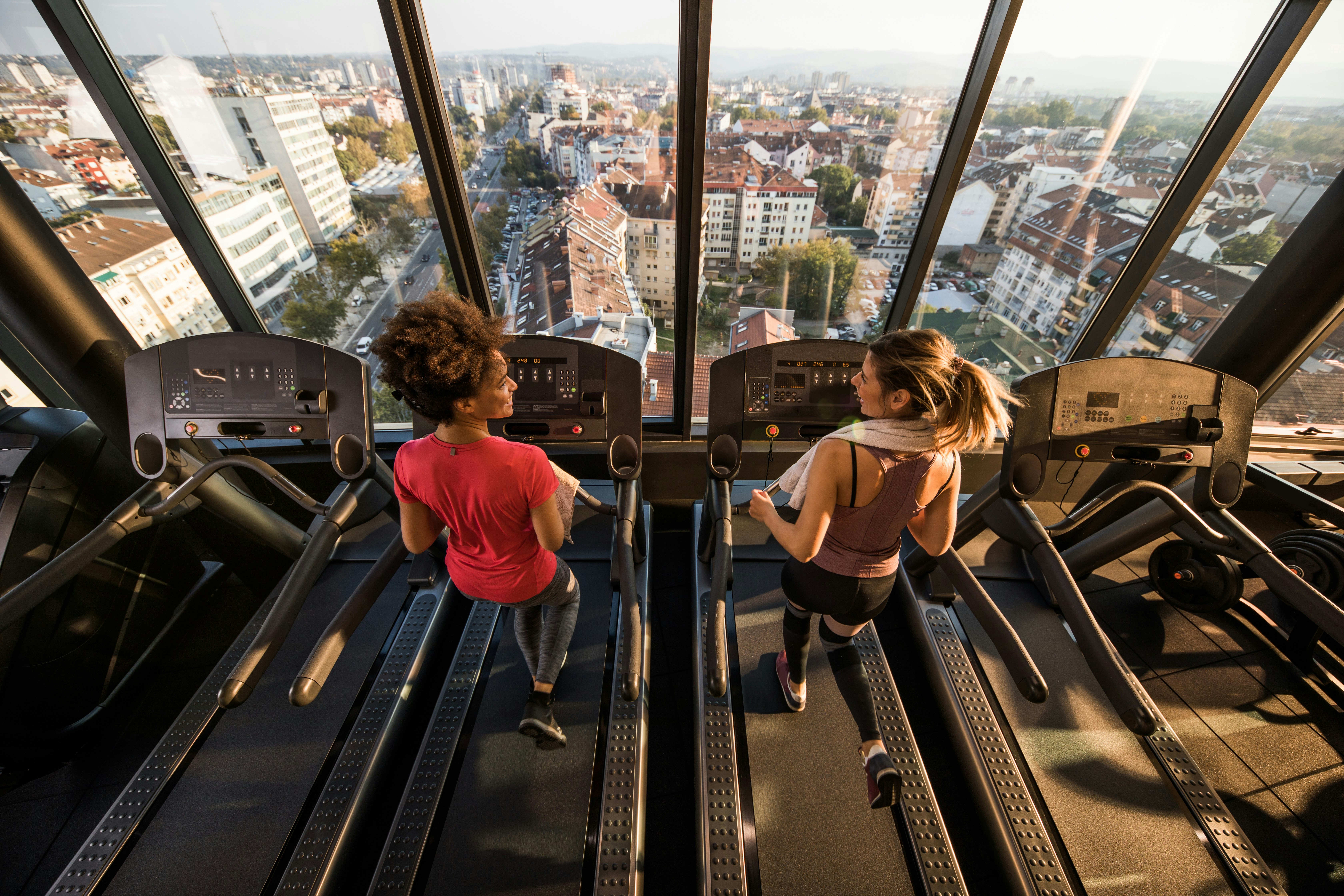 Two women run on treadmills in front a window showing a view of a city. 