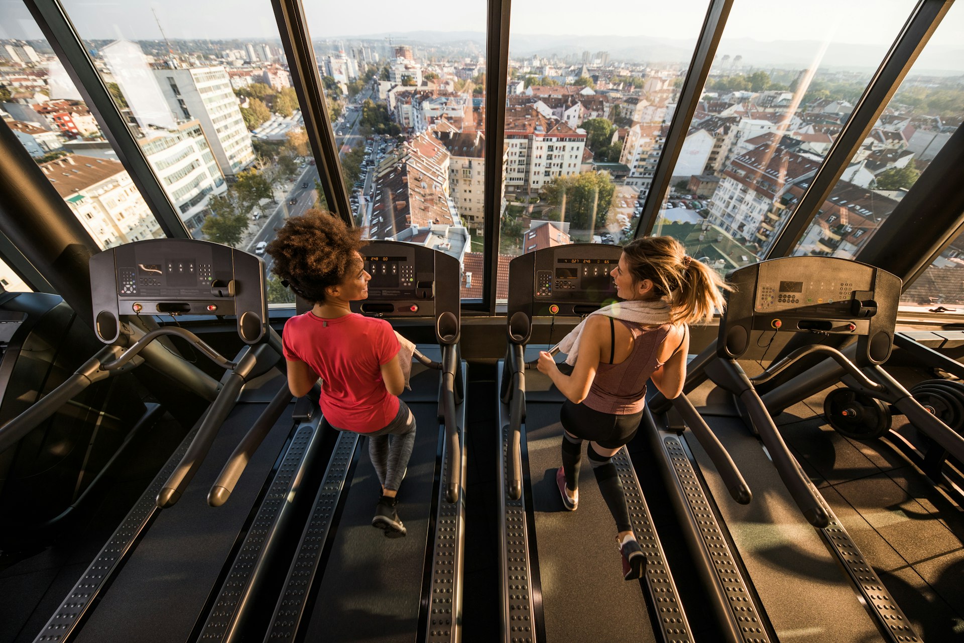 Two women run on treadmills in front a window showing a view of a city. 