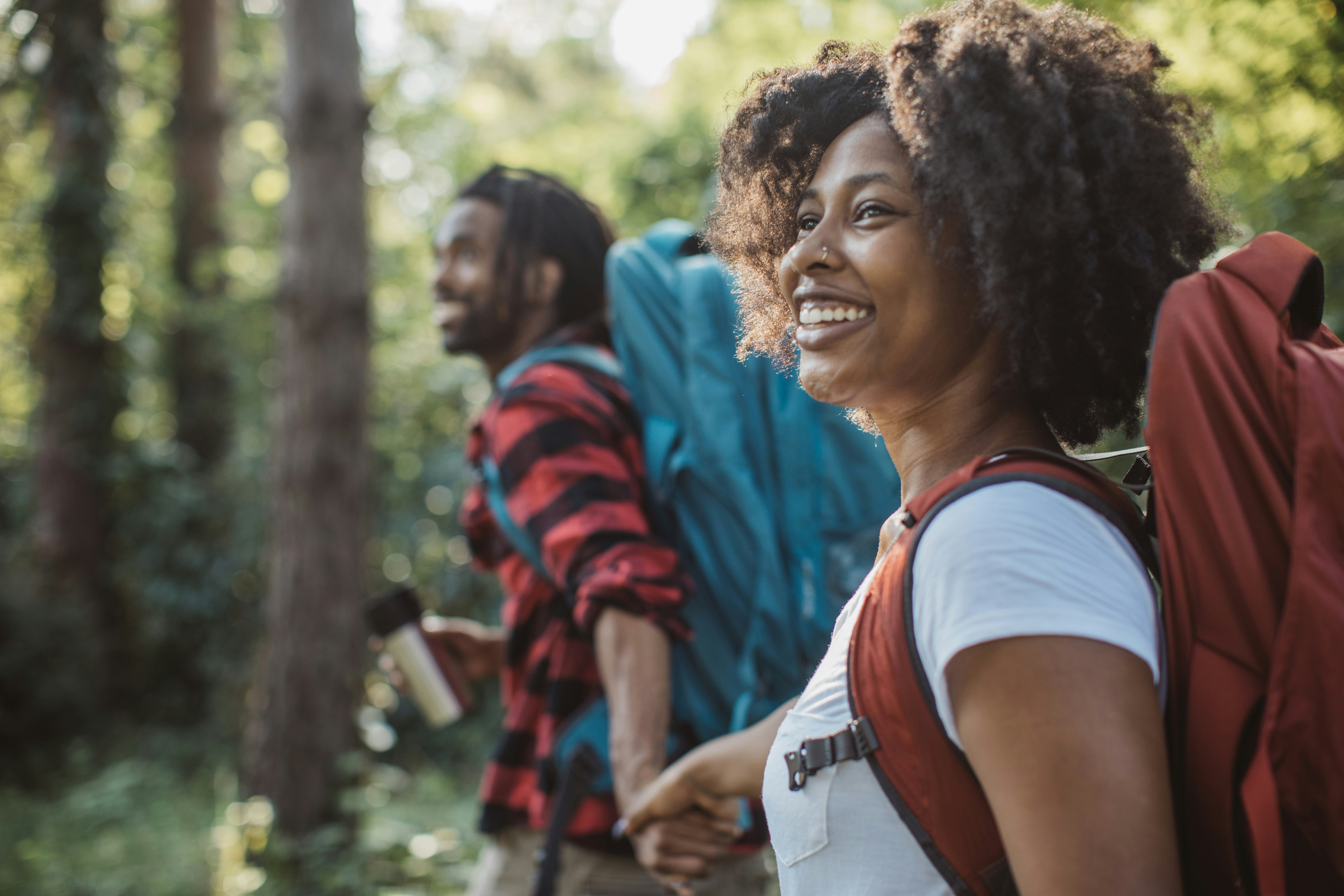 A man with locs, a red and black plaid flannel shirt, and a teal sixty liter backpack reaches for the hand of his hiking partner, who is smiling in the distance while wearing a white t-shirt and a reddish orange backpack