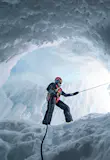 This image looks up a length of rope to a bearded man standing in an ice cave, with the taught rope leading up into the light within the cave.