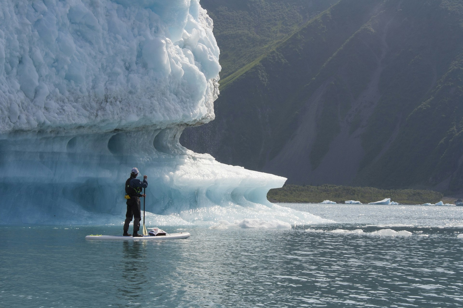 A man stands on a paddleboard in still water. A huge iceberg, white with a blueish tinge, towers over him.