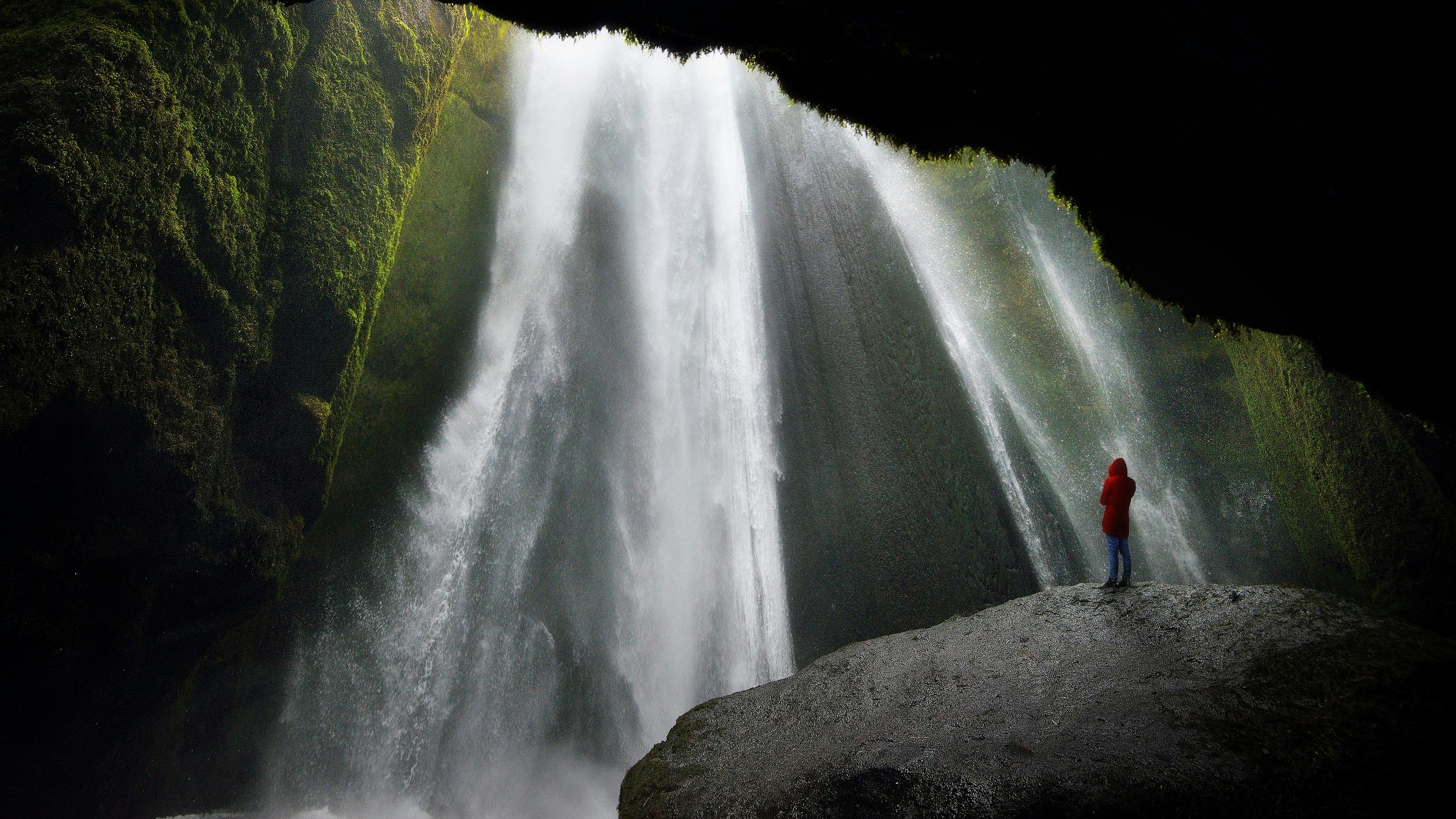 A lone person in a red jacket stands on a massive rock and stares up to a thundering waterfall crashing down in front of them; the walls of the cave are covered in vegetation.