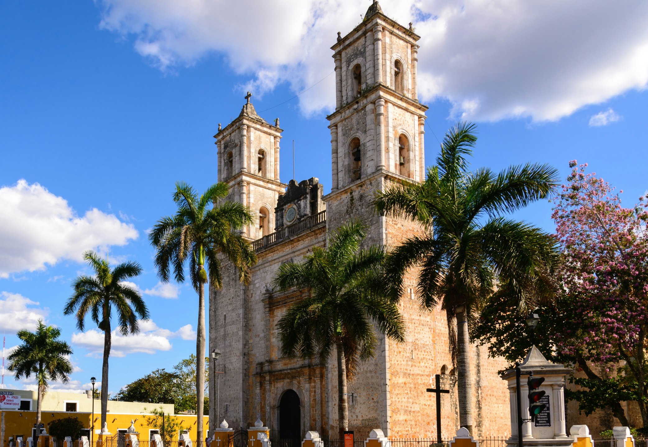 The Catedral de San Servasio in the plaza mayor of Vallodolid Mexico consists of two square stone towers on either side of a front facade with an arch shaped door in the center. The towers have two arch shaped windows on each side. Palm trees grow on the street in front of the church against a blue sky.  