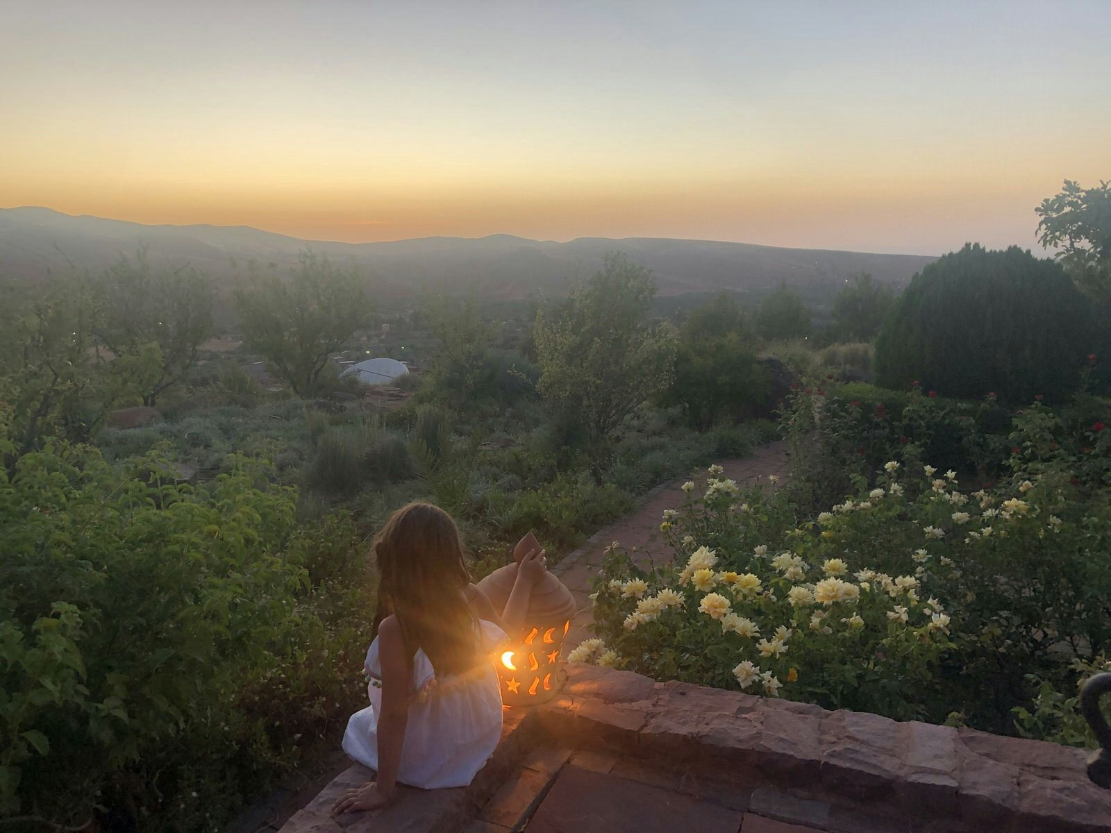 A young girl sits next to a lantern and looks out over a hazy landscape to the sunset in the distance.