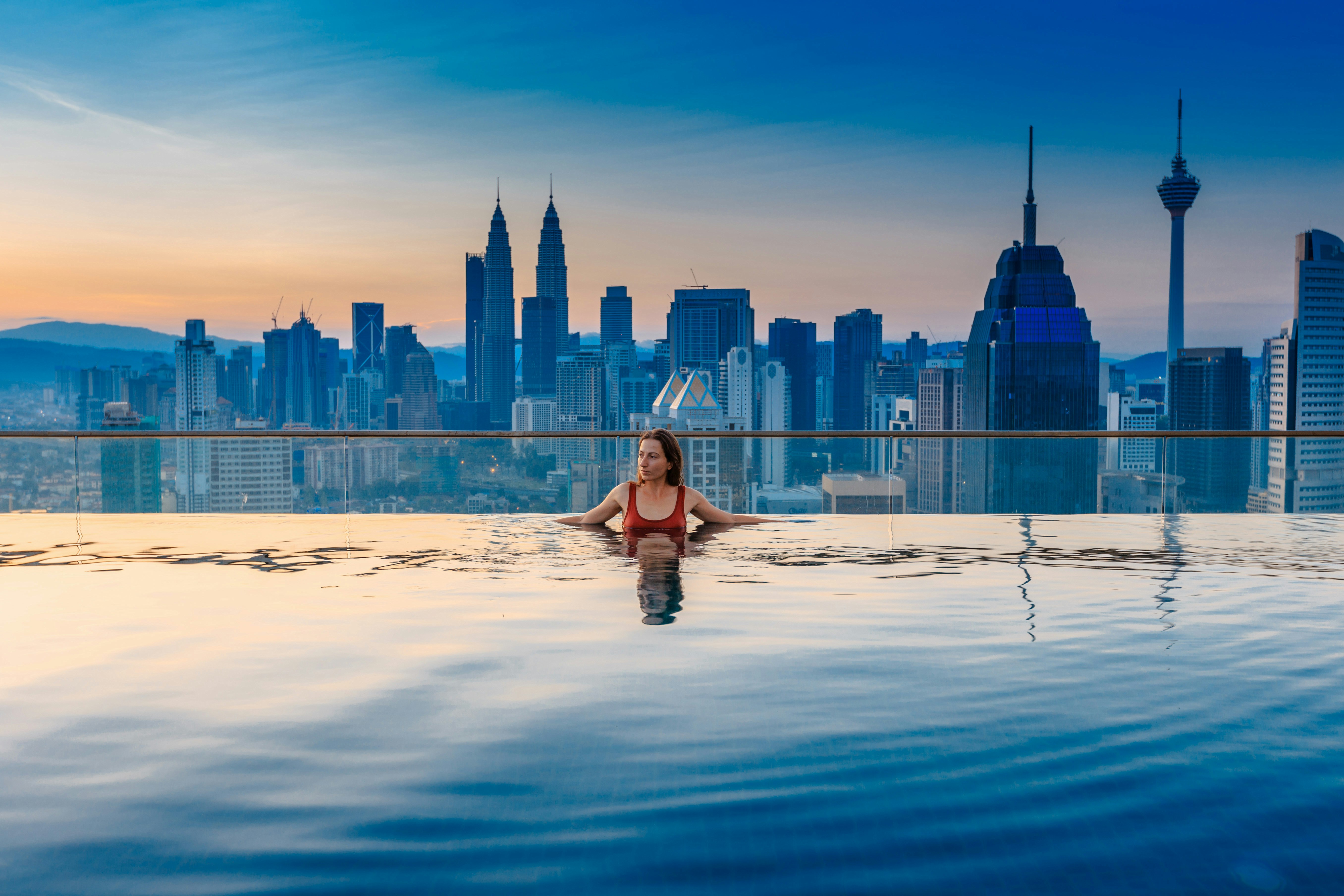 A woman in a red bathing suit swims in an infinity pool in front of a city skyline. 