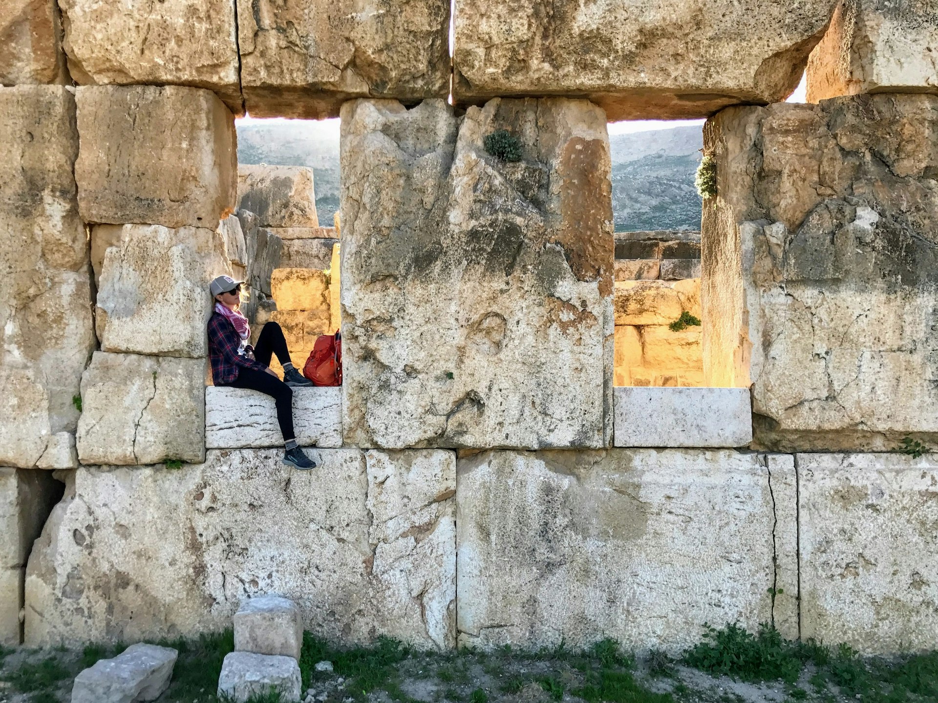 A woman sits in one of two huge open window spaces in an ancient ruin; the blocks that make up the wall are massive, each dwarfing the woman.