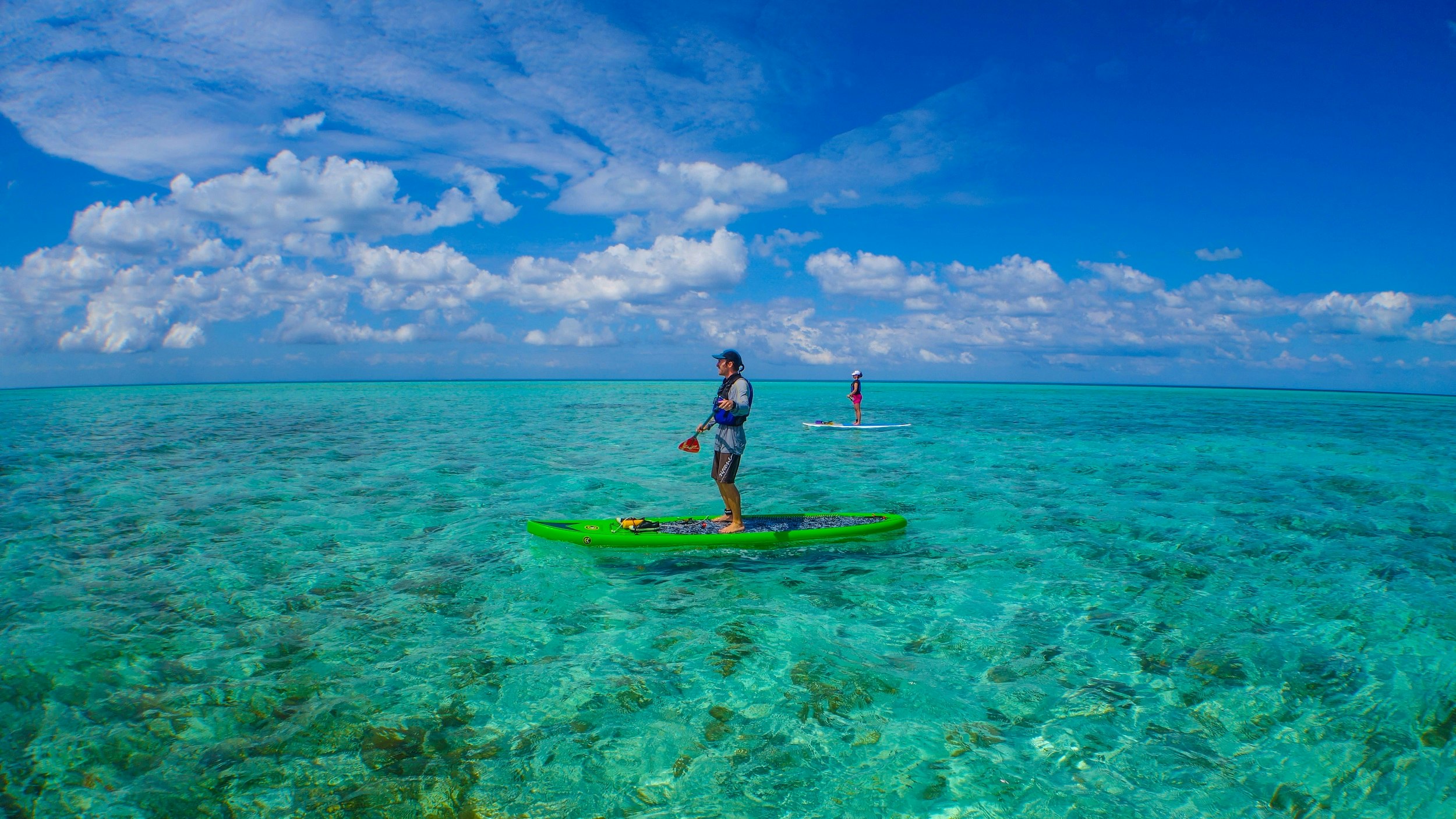 Two people, separated by 20-30m, both stand on paddle boards atop azure-coloured water, with blue skies above.