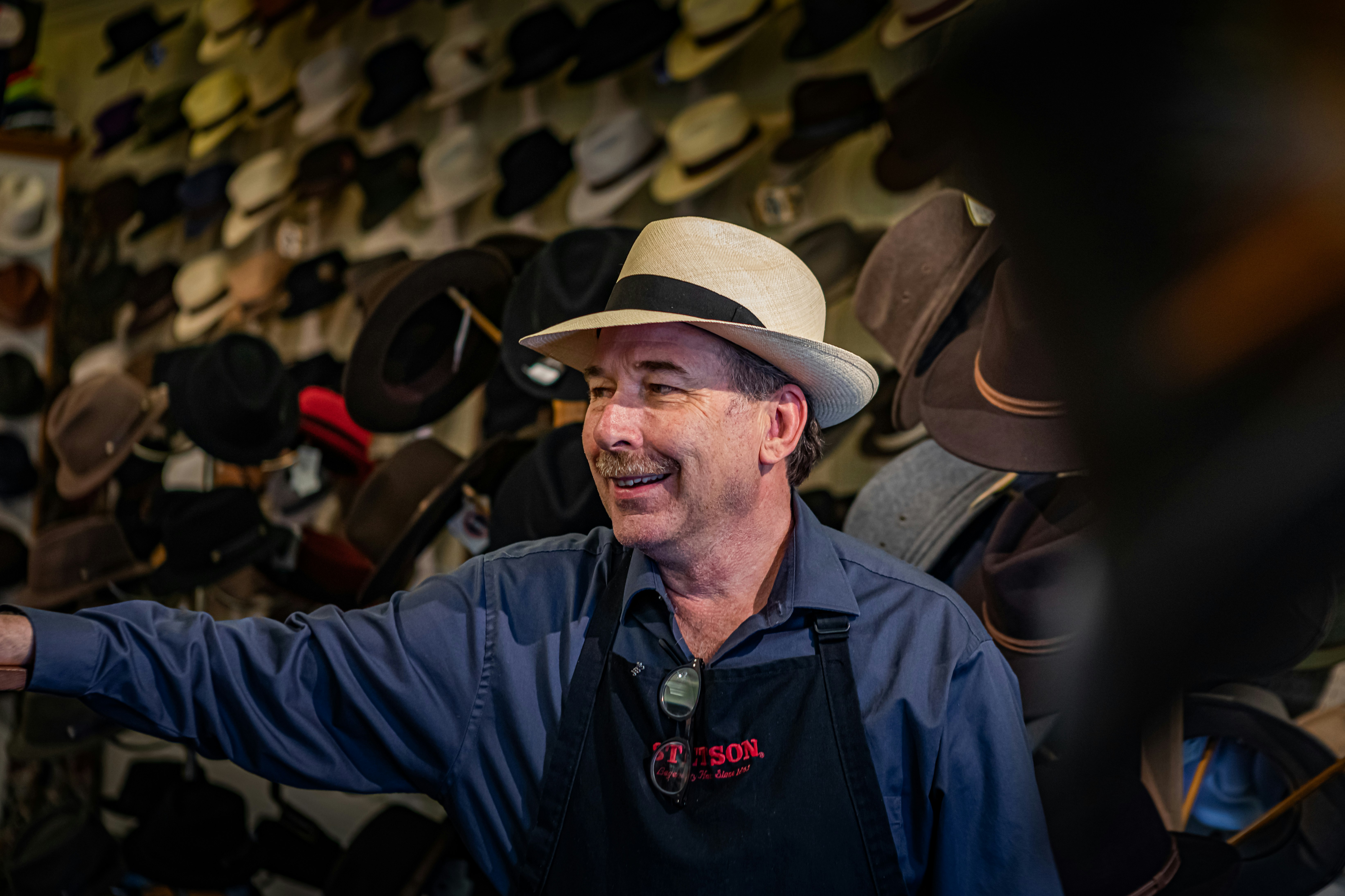 Jack Kellog, owner of Hatman Jack's in Wichita, stands in a black apron and dark button down shirt with an easy, comfortable smile in front of a wall of hats hung on pegs. 