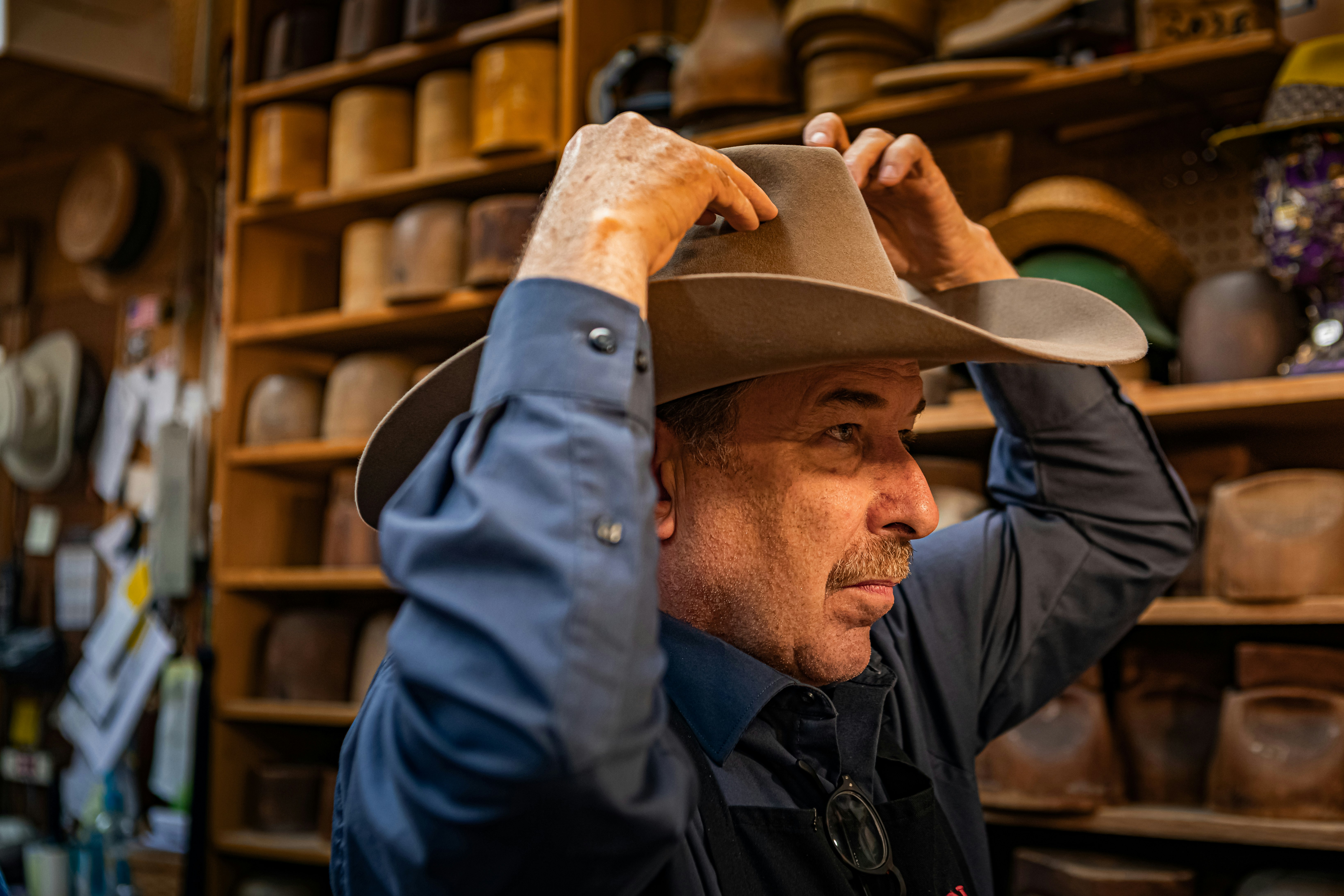 Jack Kellogg stands in profile to the viewer as he adjusts a large Stetson cowboy hat in a light brown color. In the background are many wooden hat molds on shelves