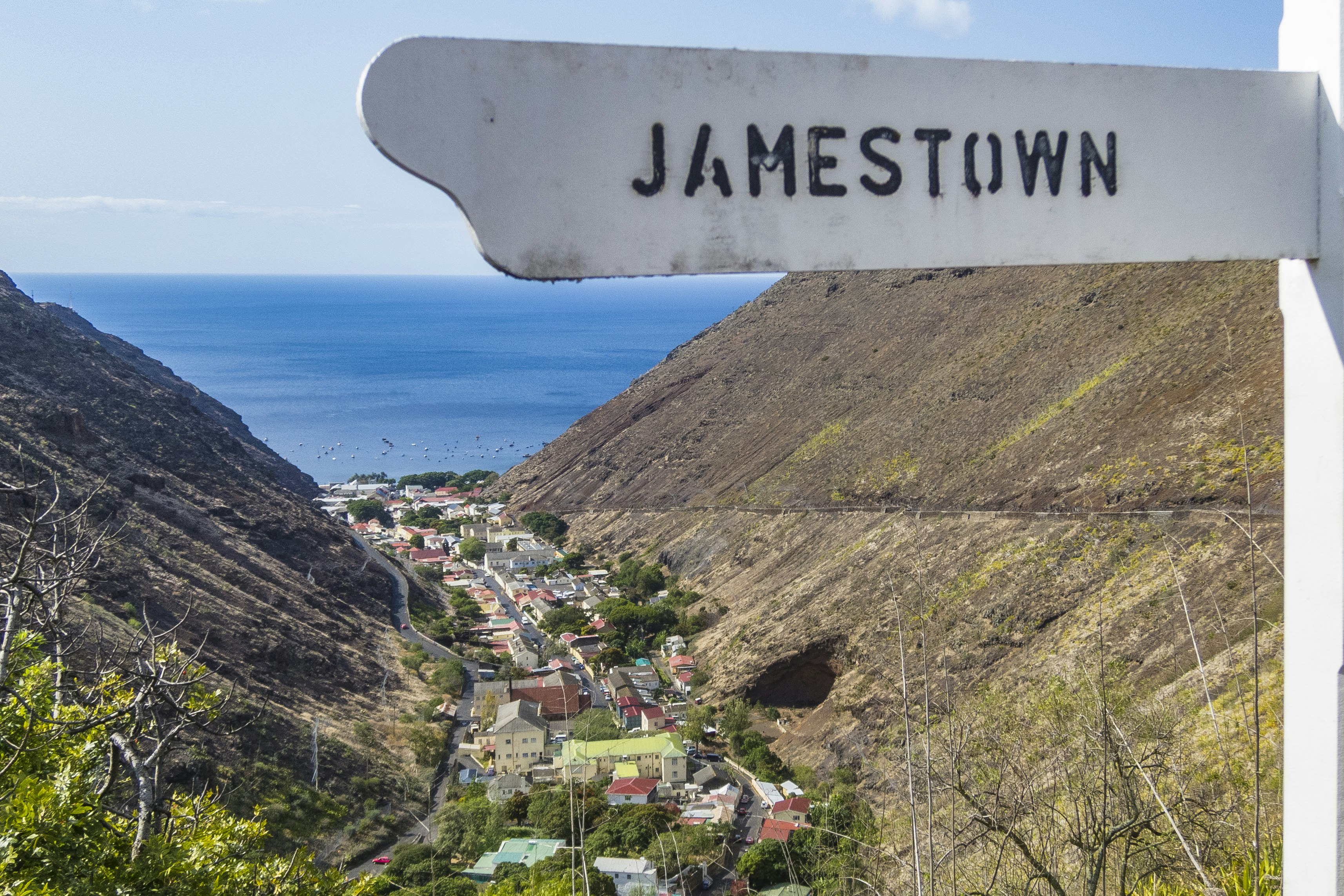 A cute hand-like white street sign reading 'Jamestown' hangs from a pole; the horizon on the Atlantic Ocean sits just below the sign, and below that is the town of Jamestown nestled between two steep slopes.