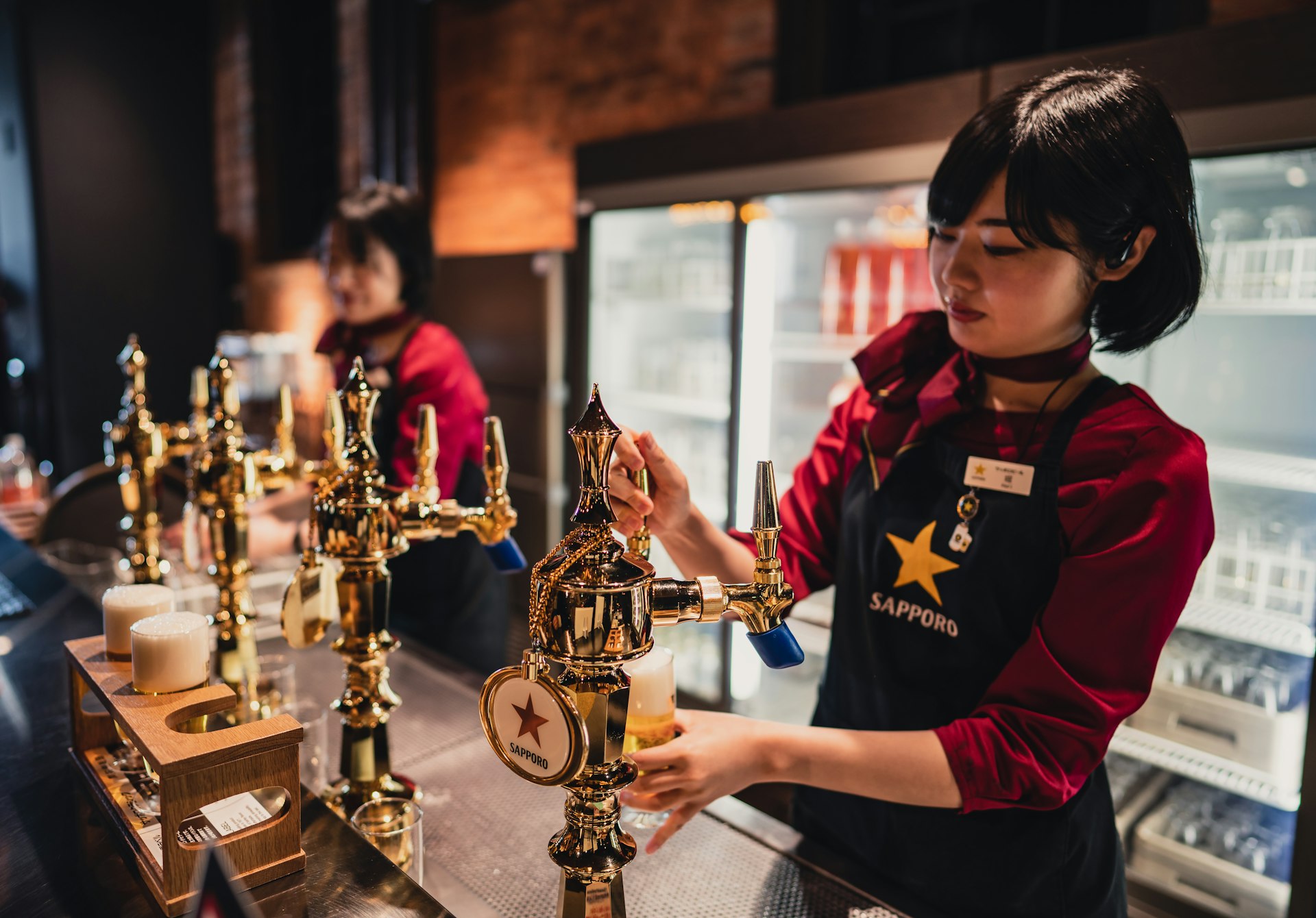A woman with a black bob haircut in a red shirt and a black apron with a yellow star and the Sapporo logo pours a beer from a vintage-looking brass tap in a bar full of natural light at the Sapporo Beer Museum