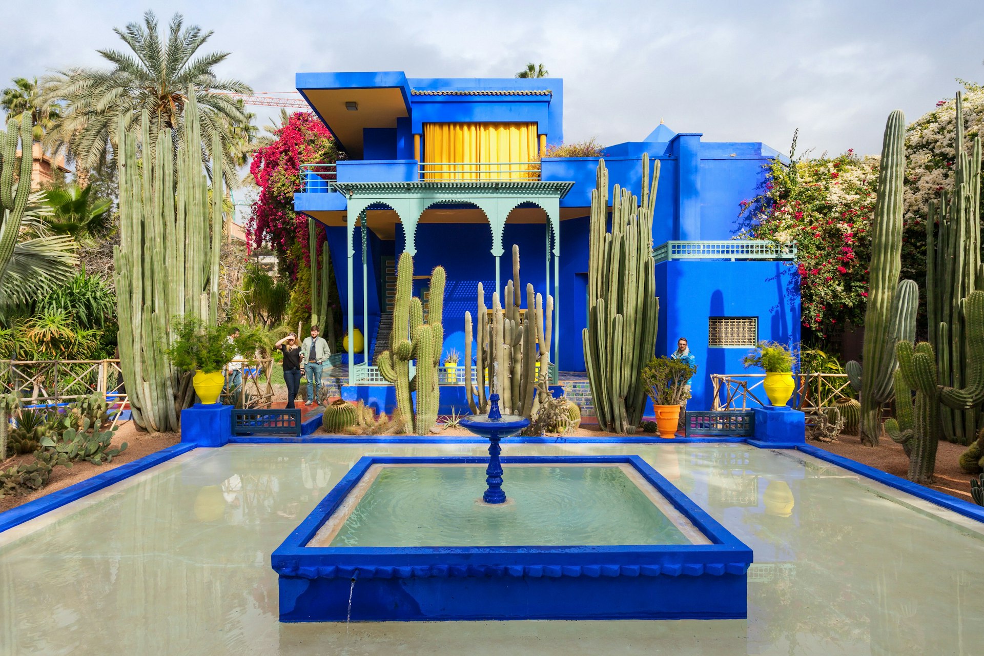 A bright blue fountain sits in a small blue pool, which is inset in a larger blue-trimmed pool; in the background, surrounded by cacti and palms is an art-deco villa in the same colour blue (but with gold curtains).