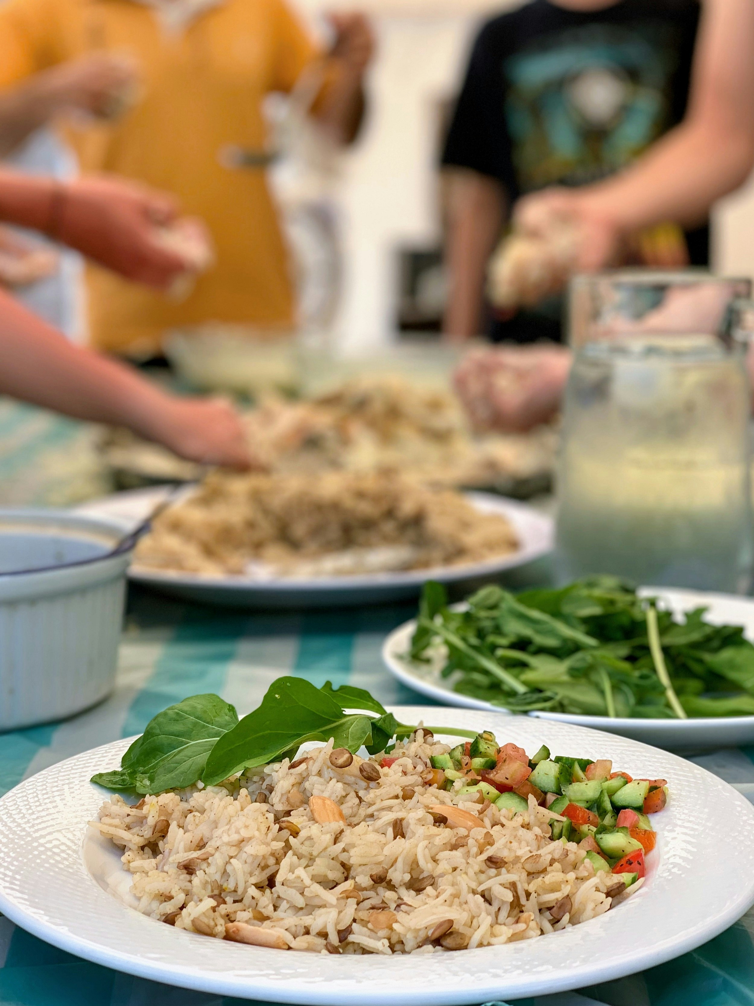 A shot looking down a table at plates filled with a rice and lentil dish; the plate nearest the camera is in focus, while all the others are blurred (along with various people's arms extending over the table at the far end).