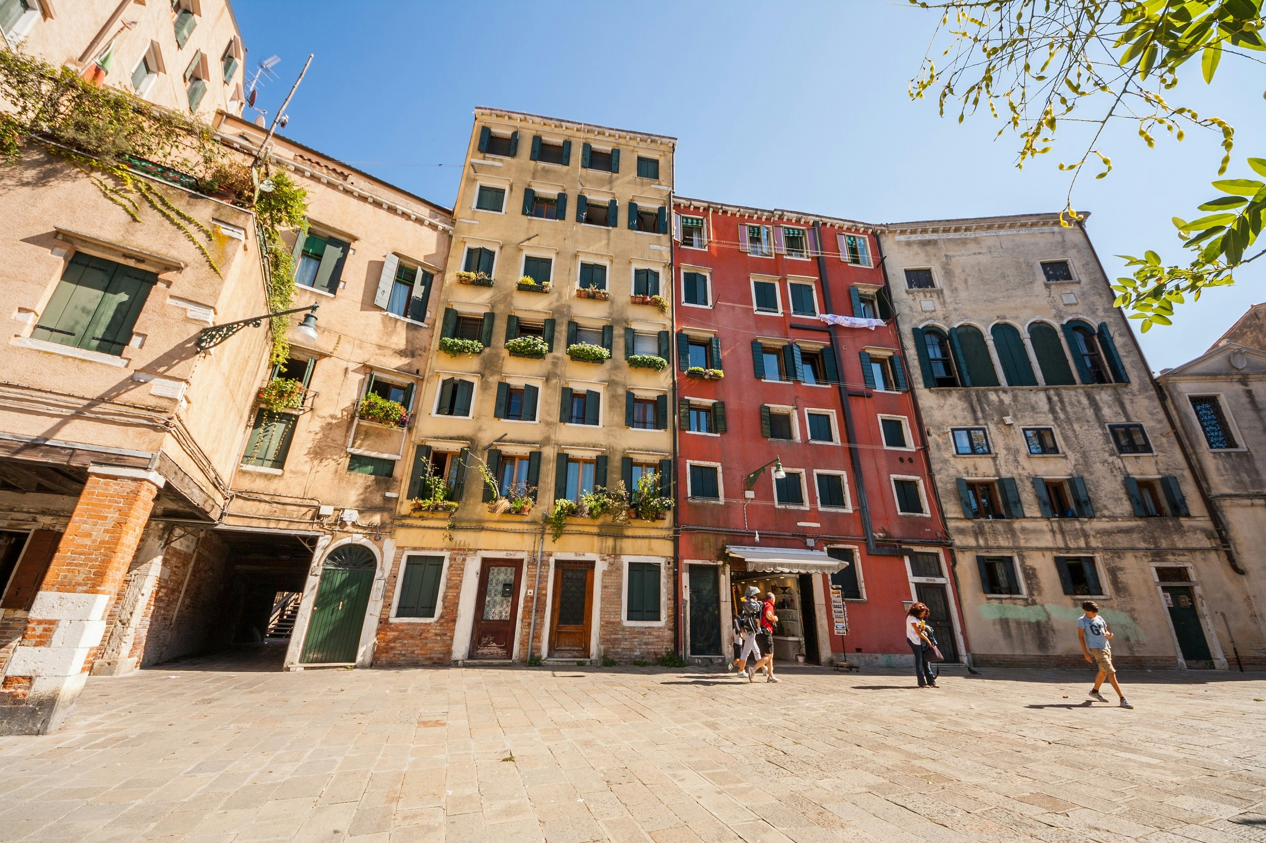 An open area dominated by three six-storey buildings with pastel-coloured facades. Four people can be seen strolling by