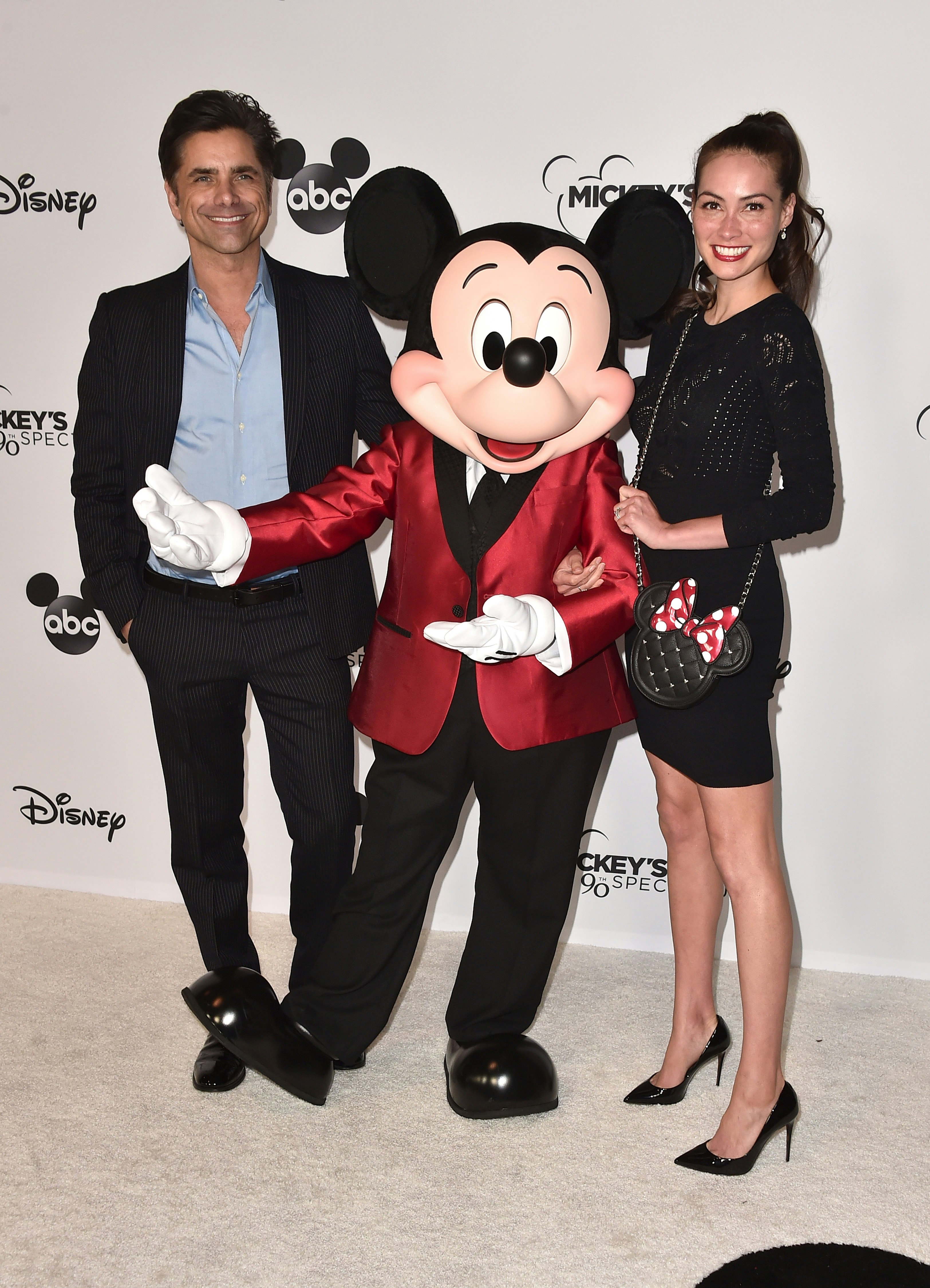 John Stamos and Caitlin McHugh posing with Mickey Mouse in front of a backdrop of Disney logos.