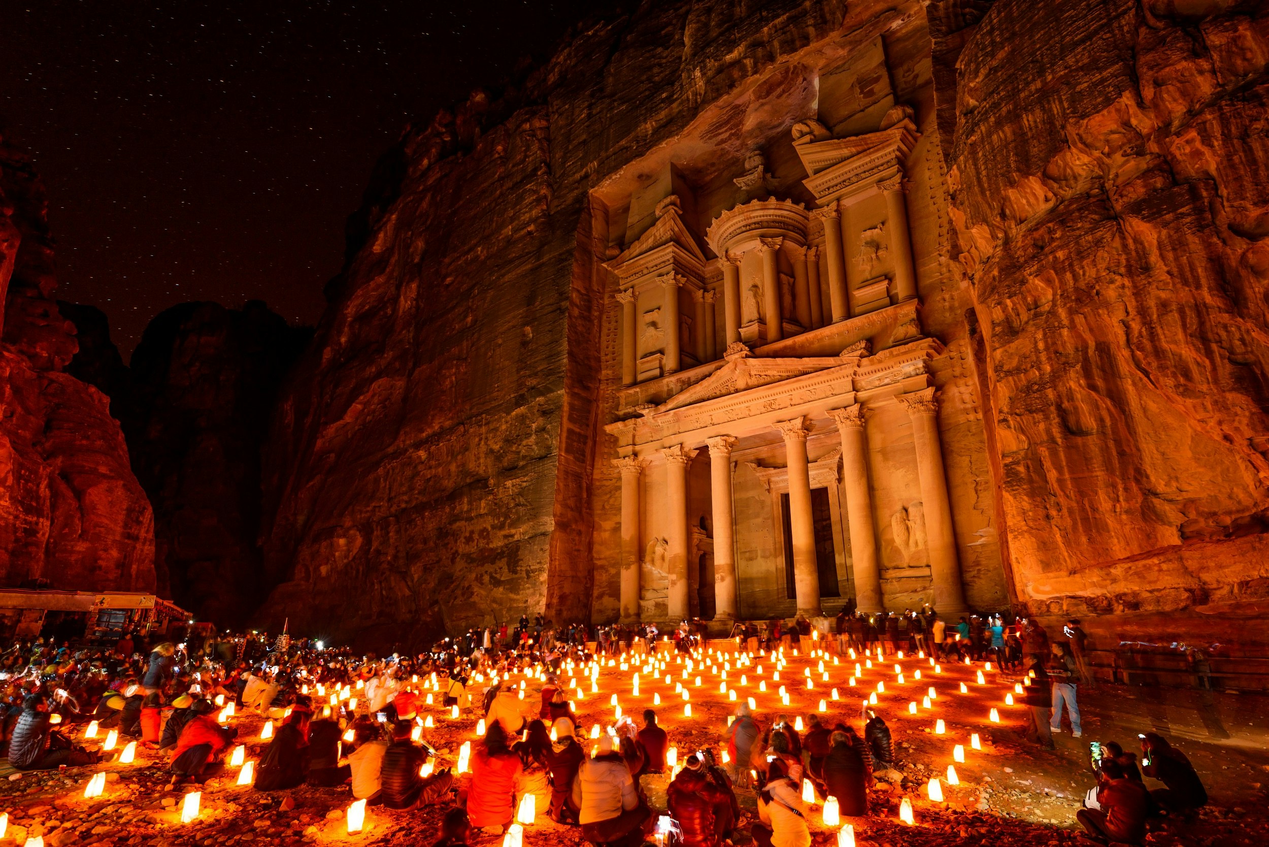 The intricate facade of the Treasury at Petra, which was carved into a cliff centuries ago; the whole seen is glowing from candles on the ground, which sit amongst a crowd of visitors.