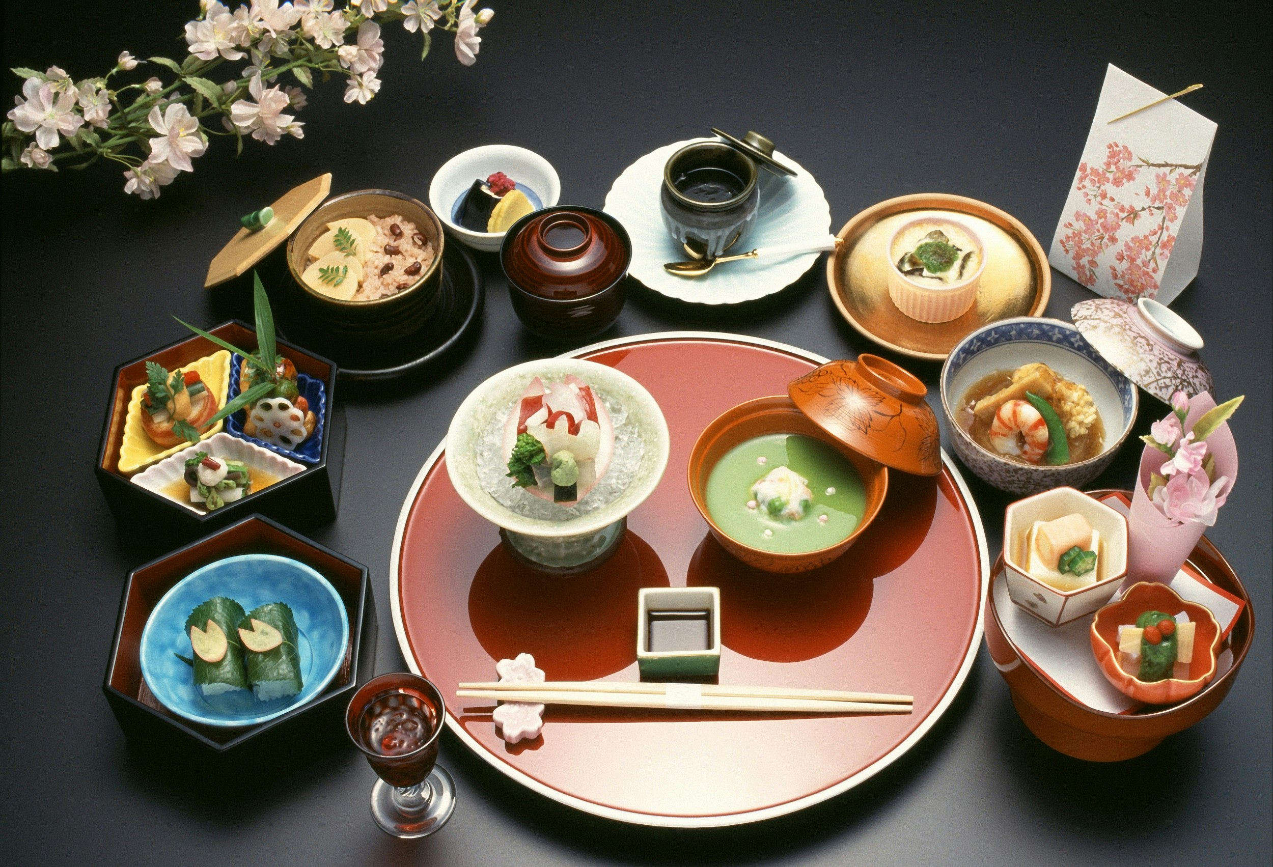 A traditional kaiseki meal sits on a dark table; there are two bowls full of ornate looking dishes atop a red plate, and surrounding it are nine bowls and dishes of various shapes and sizes, each with delicate morsels of food.