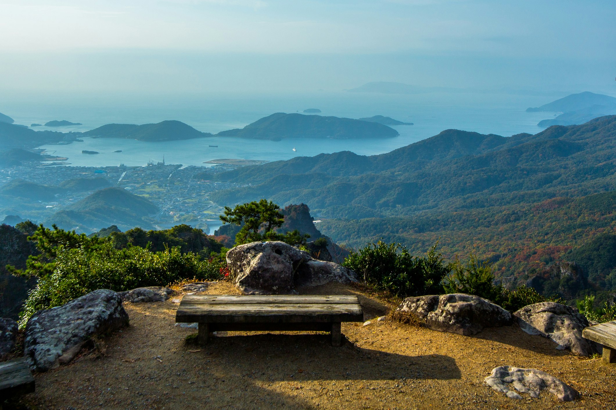 A low wooden bench sits empty on an overlook with stellar views of the Kankakei gorge. In the distance, low islands rise out of the denim-colored sea, which blends into the hazy sky