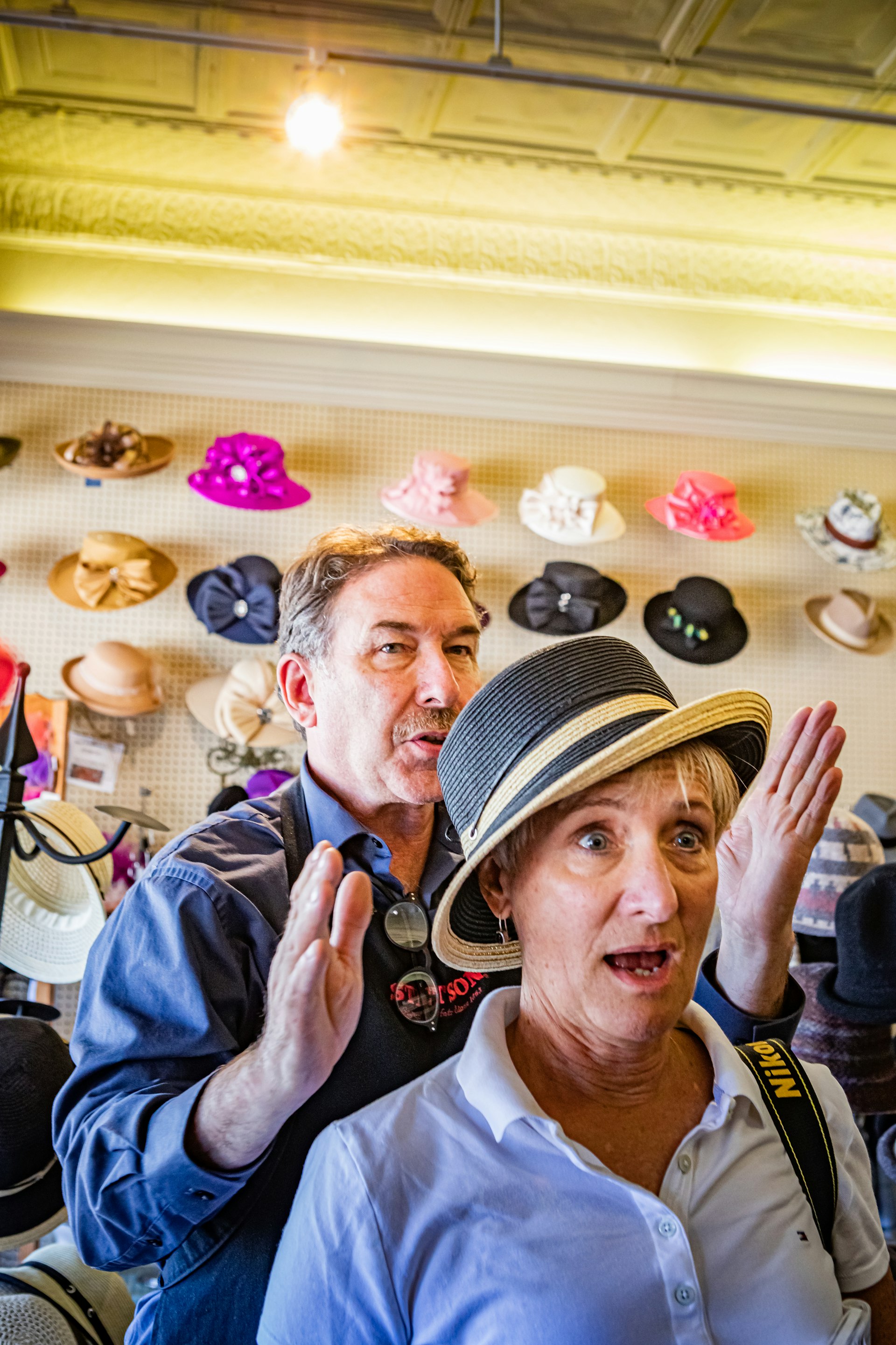 Jack Kellogg stands behind a female customer with short blond hair as she tries on a black and cream straw hat. He has a look of confident, kind expertise on his face and is gesturing as if they've found just the right angle. The customer's face is one of pleased surprise as she sees how the hat accentuates her facial features.