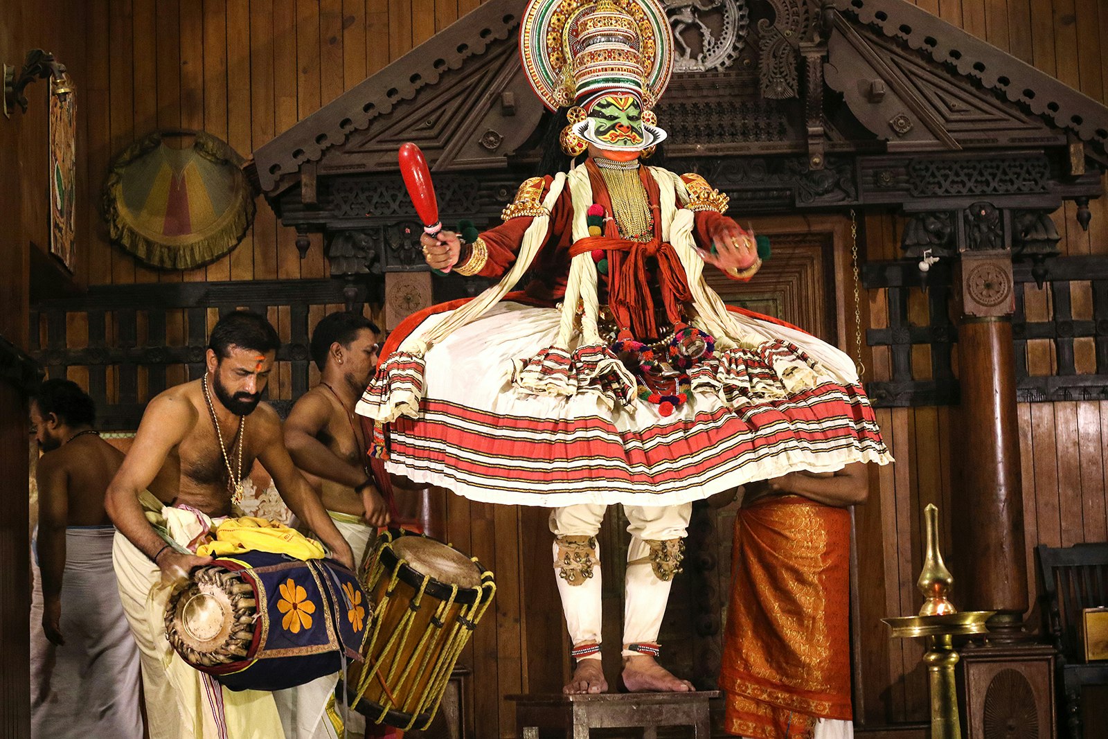 A Kathakali performer dancers on a small table while two men play drums behind him. His face is painted green and he wears a headdress, and a red and white skirt. Kochi, India.