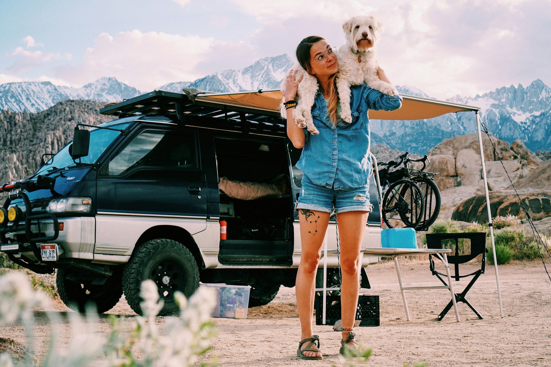 Katie Boue stands in front of her van with dog Spaghetti around her shoulders. She is wearing a cambray shirt, jean shorts, and round glasses. Between her and the van is a pop-up shade canopy and foldable camping chairs.