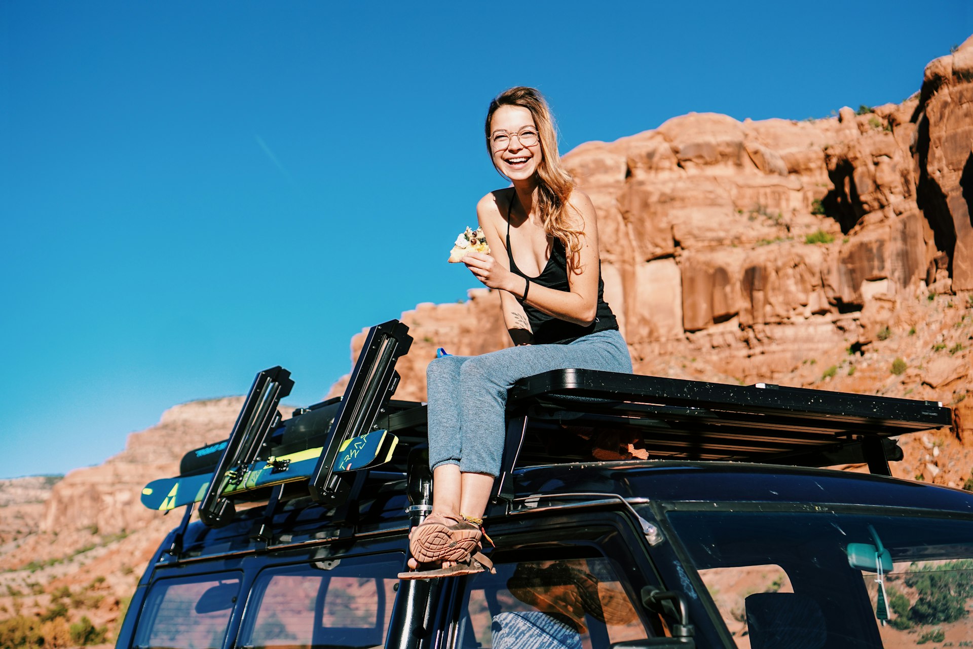 Katie_Boué sits on top of her van's roof rack in grey athletic pants and a black tank top, smiling with her hair in a pony tail and round glasses on her face. She is holding a slice of pizza. 