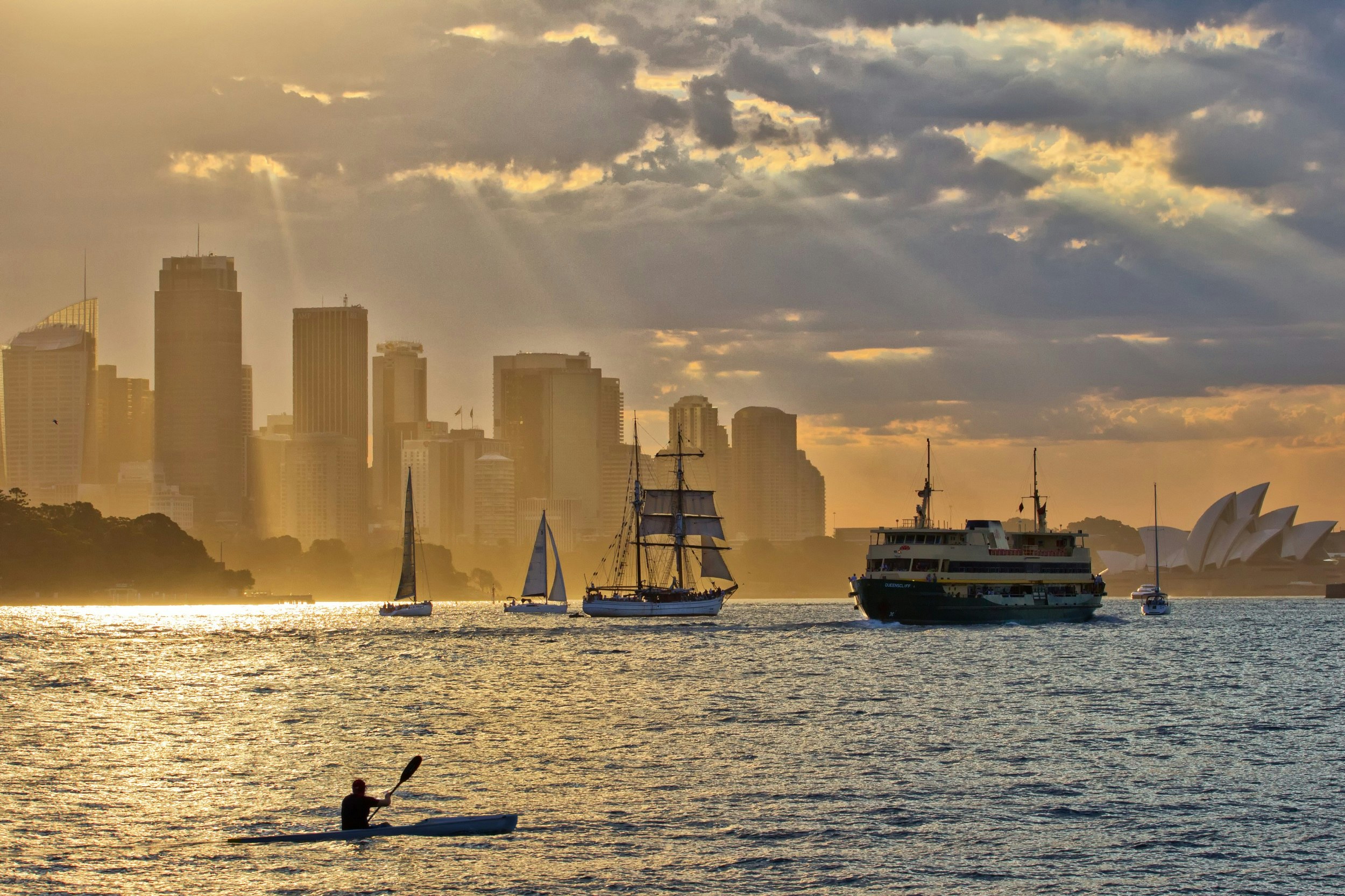 A lone man paddles a surfski across the waters of Sydney Harbour; the late afternoon sun pierces the dark clouds above and sends golden rays down into the high-rise buildings, the Sydney Opera House and other boats on the water (ferries, sailboats etc).