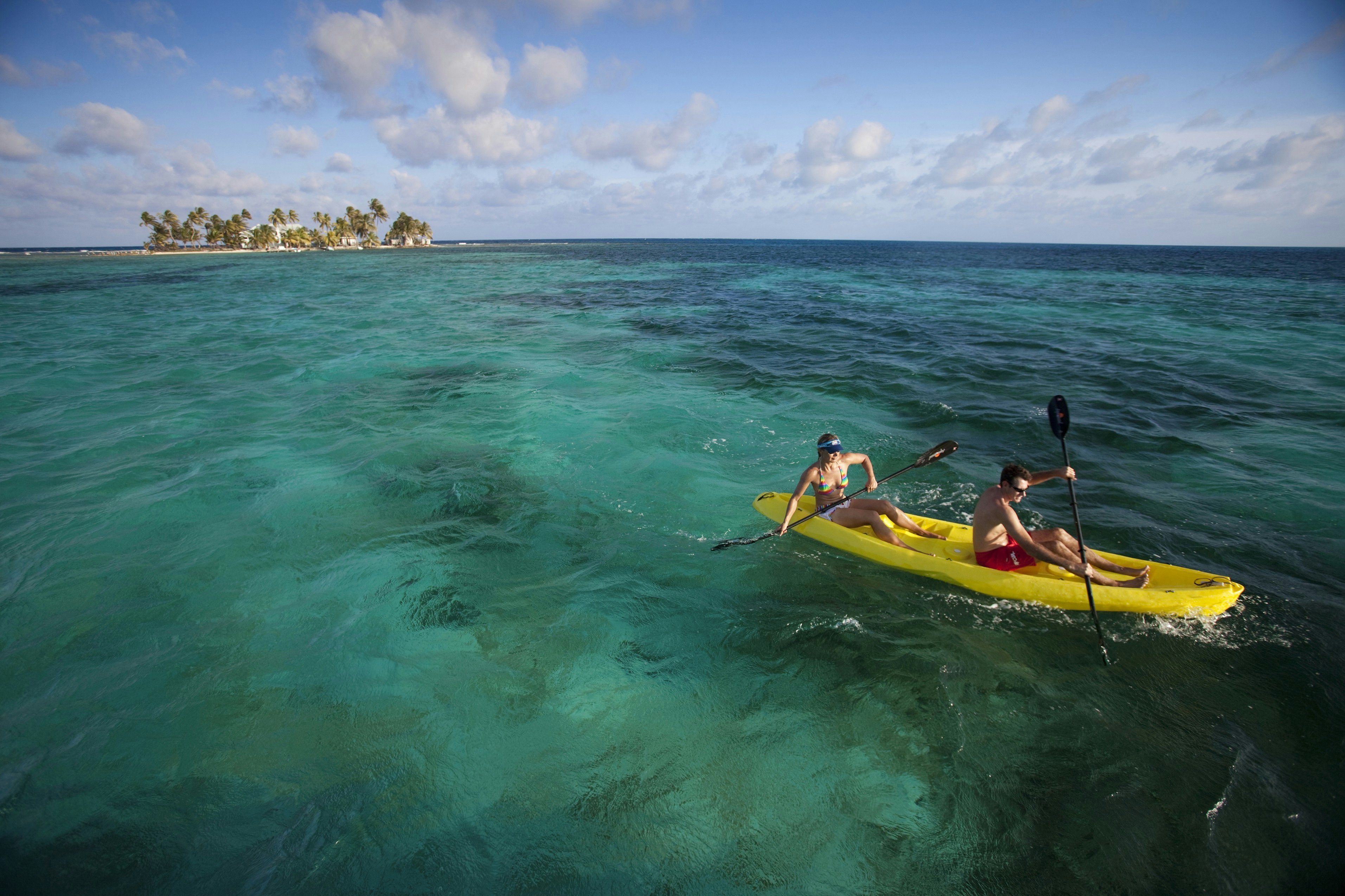 A man and woman wearing swimsuits sit in an open-topped kayak and paddle away from a small island in tropical waters
